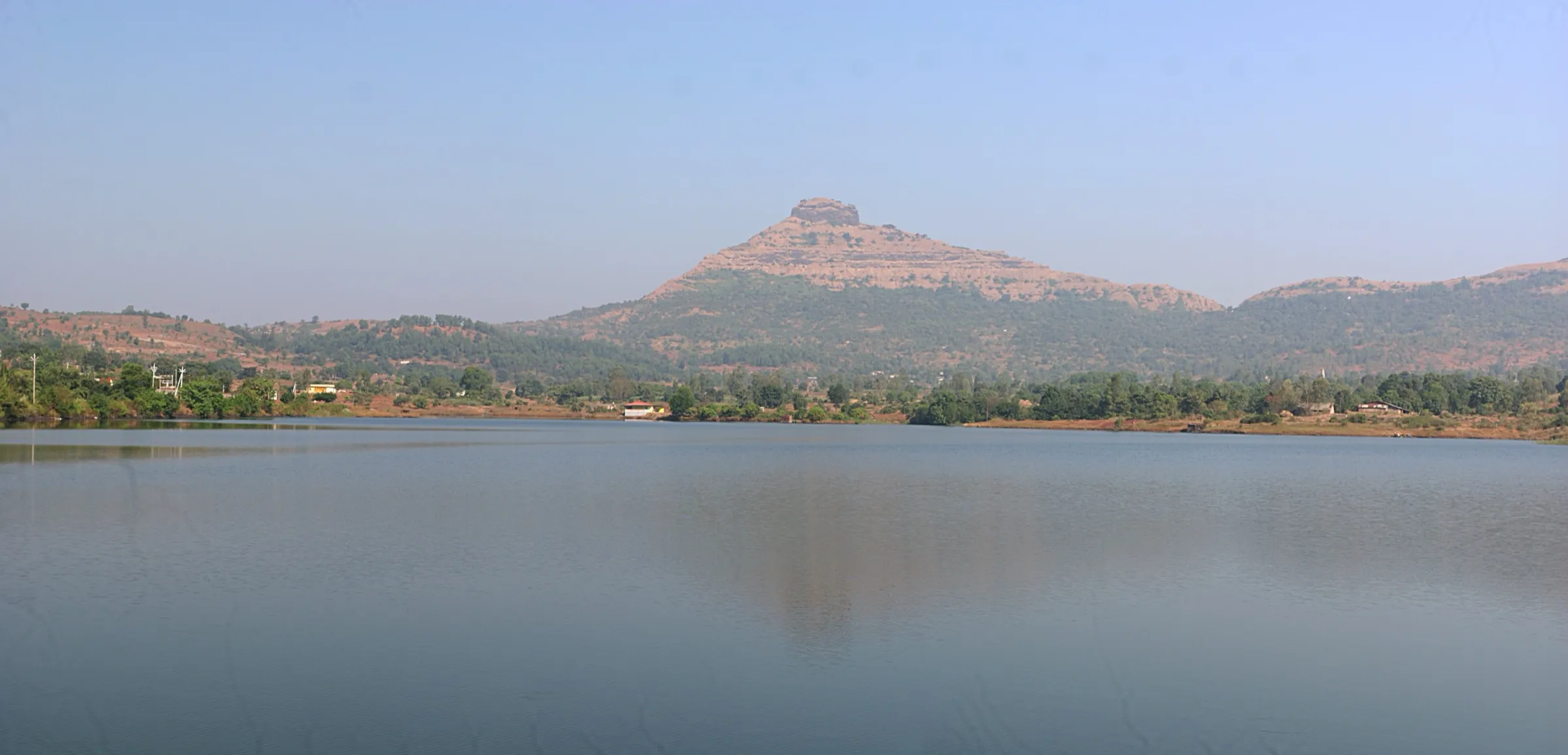 The pyramid-shaped Tikona Fort hill seen across the still water of Hadashi dam under a clear sky