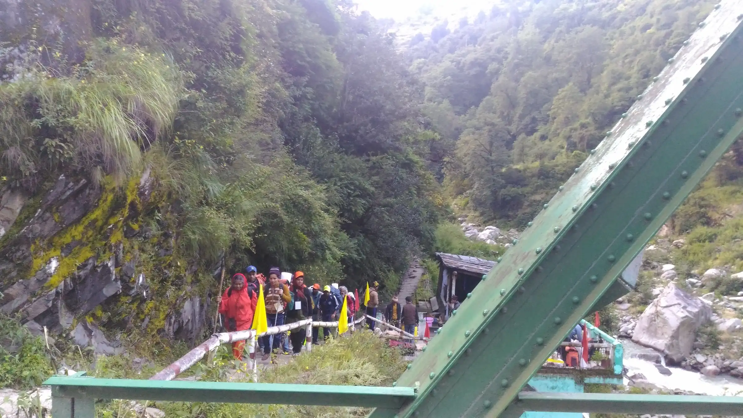Trekkers carrying yellow flags walk off a green steel bridge over the Hanuman Ganga at Hanuman Chatti, the start of the trail