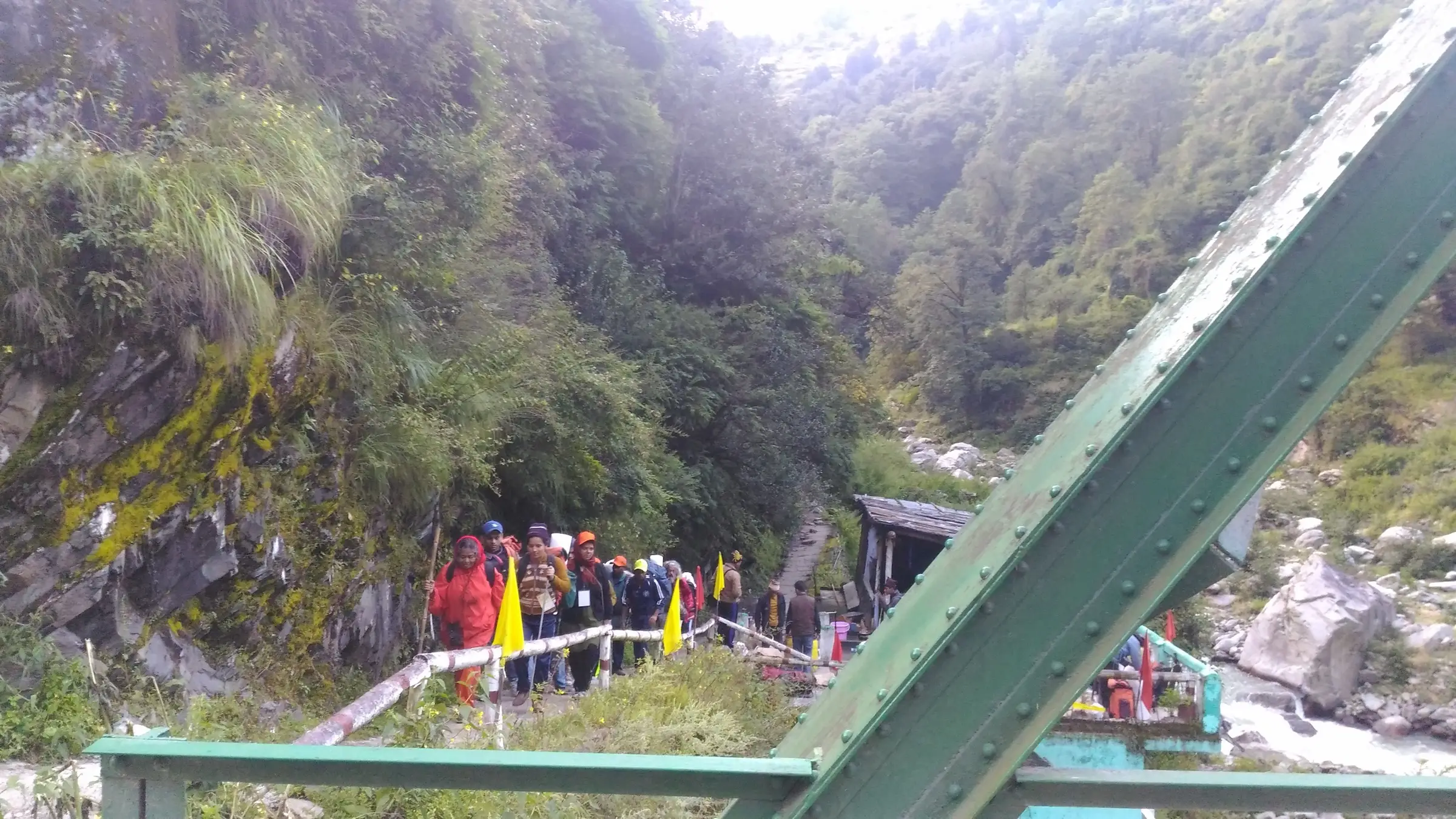 Trekkers carrying yellow flags walk off a green steel bridge over the Hanuman Ganga at Hanuman Chatti, the start of the trail