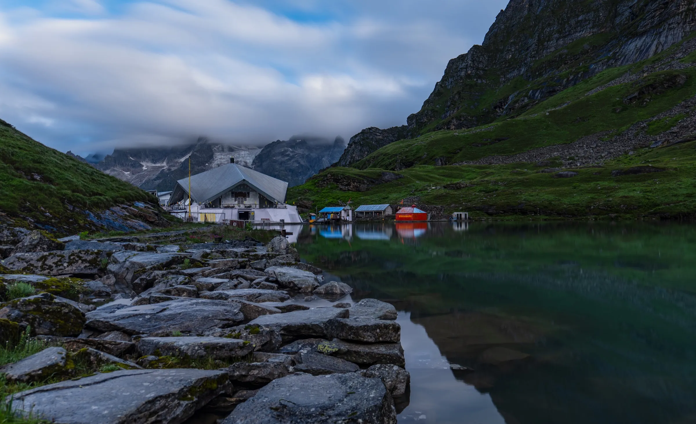 Gurudwara Shri Hemkunt Sahib and small shelters on the shore of the still Hemkund (Lokpal) lake below cloud-capped peaks