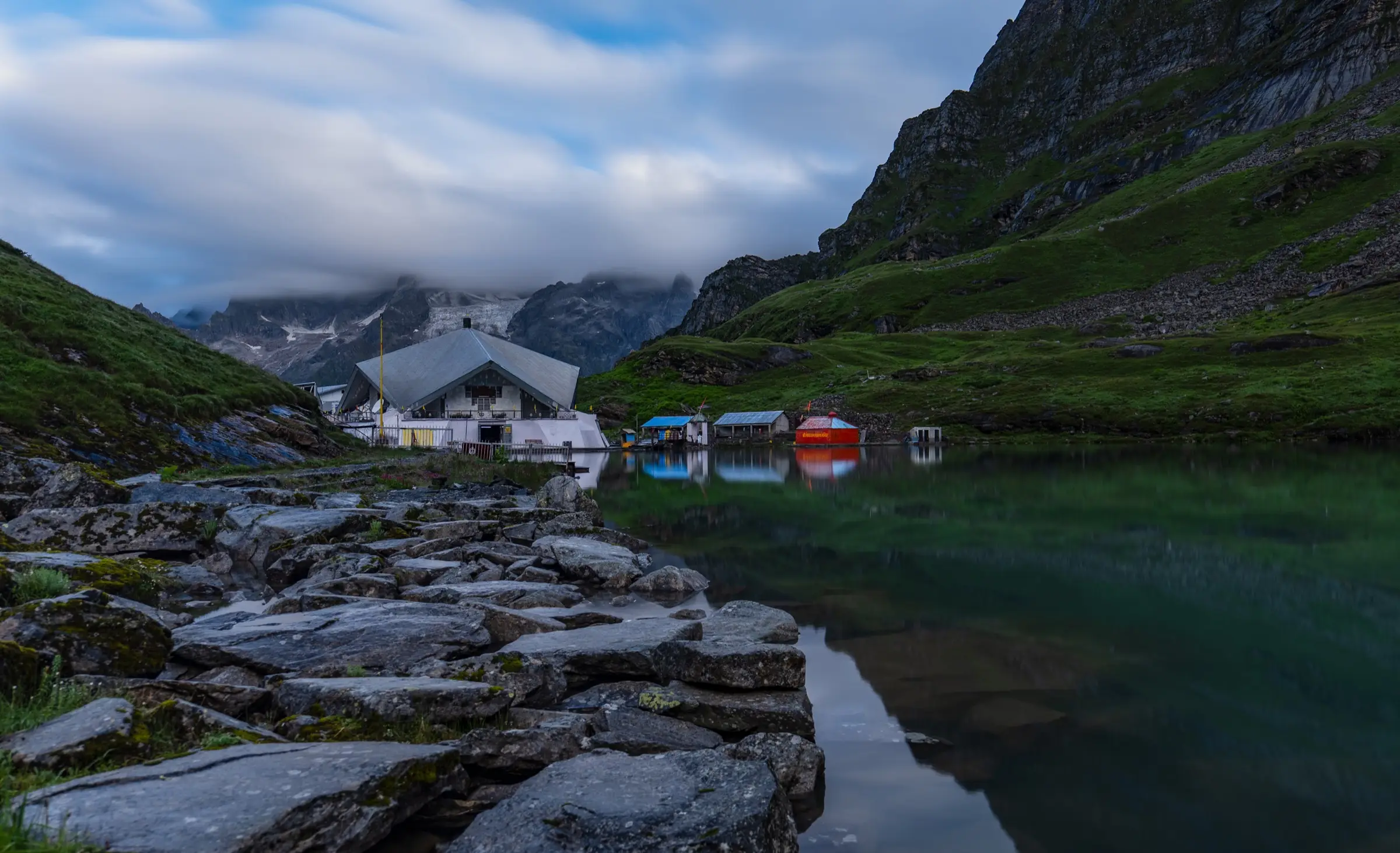 Gurudwara Shri Hemkunt Sahib and small shelters on the shore of the still Hemkund (Lokpal) lake below cloud-capped peaks