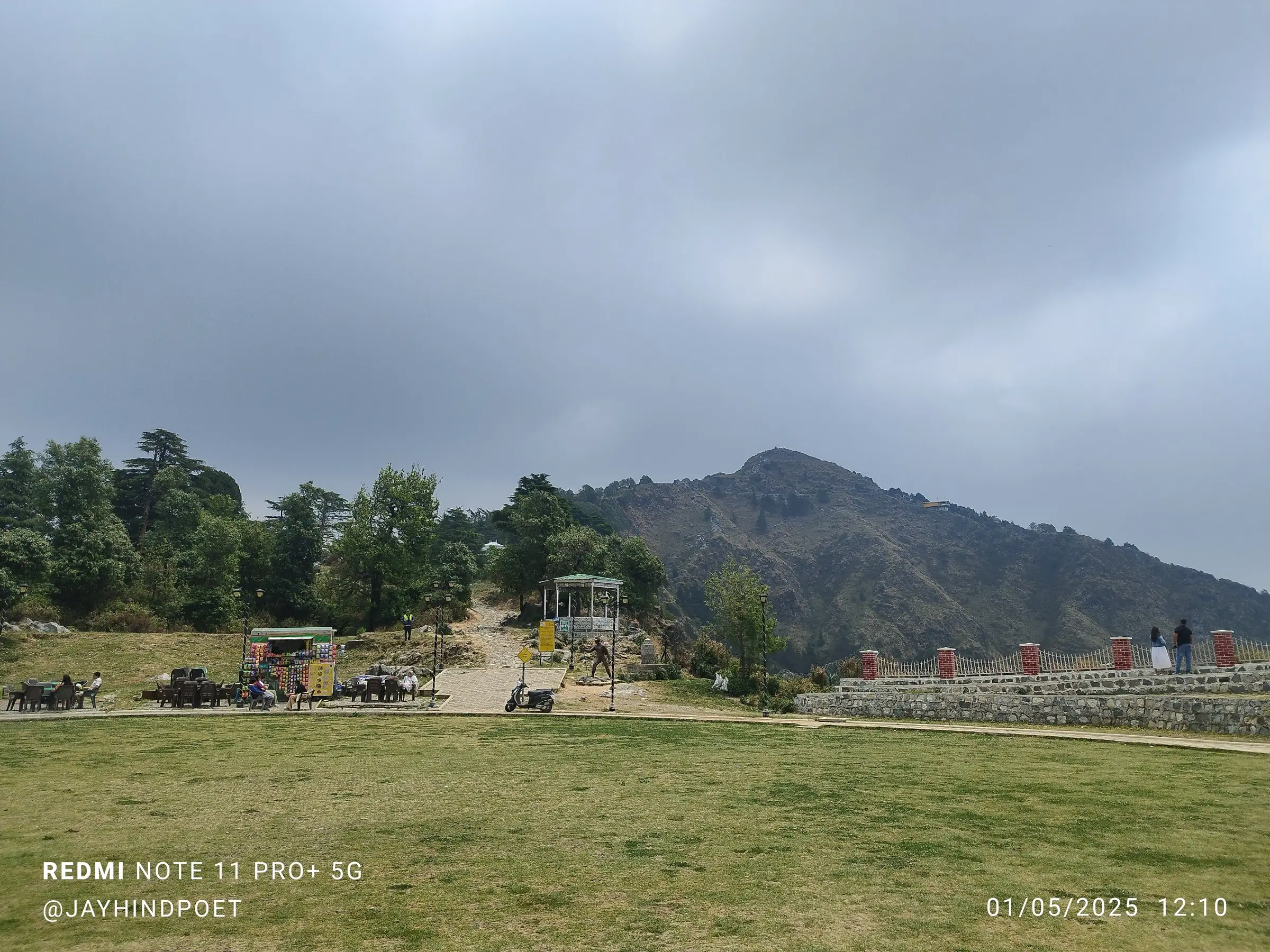 Lawn of the George Everest heritage site with food stalls and a gazebo, the stone path leading up toward the bare George Everest Peak under grey cloud