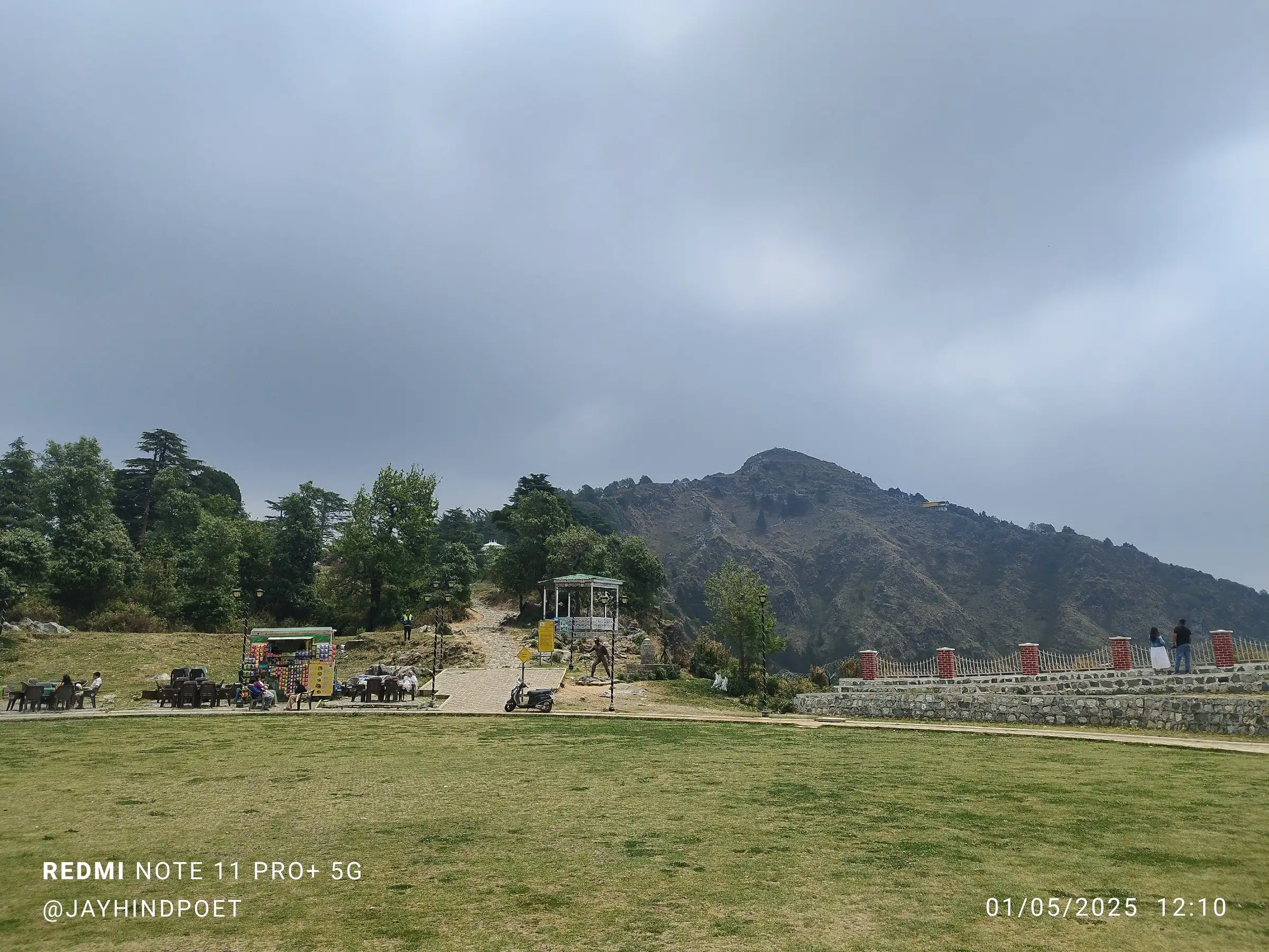 Lawn of the George Everest heritage site with food stalls and a gazebo, the stone path leading up toward the bare George Everest Peak under grey cloud