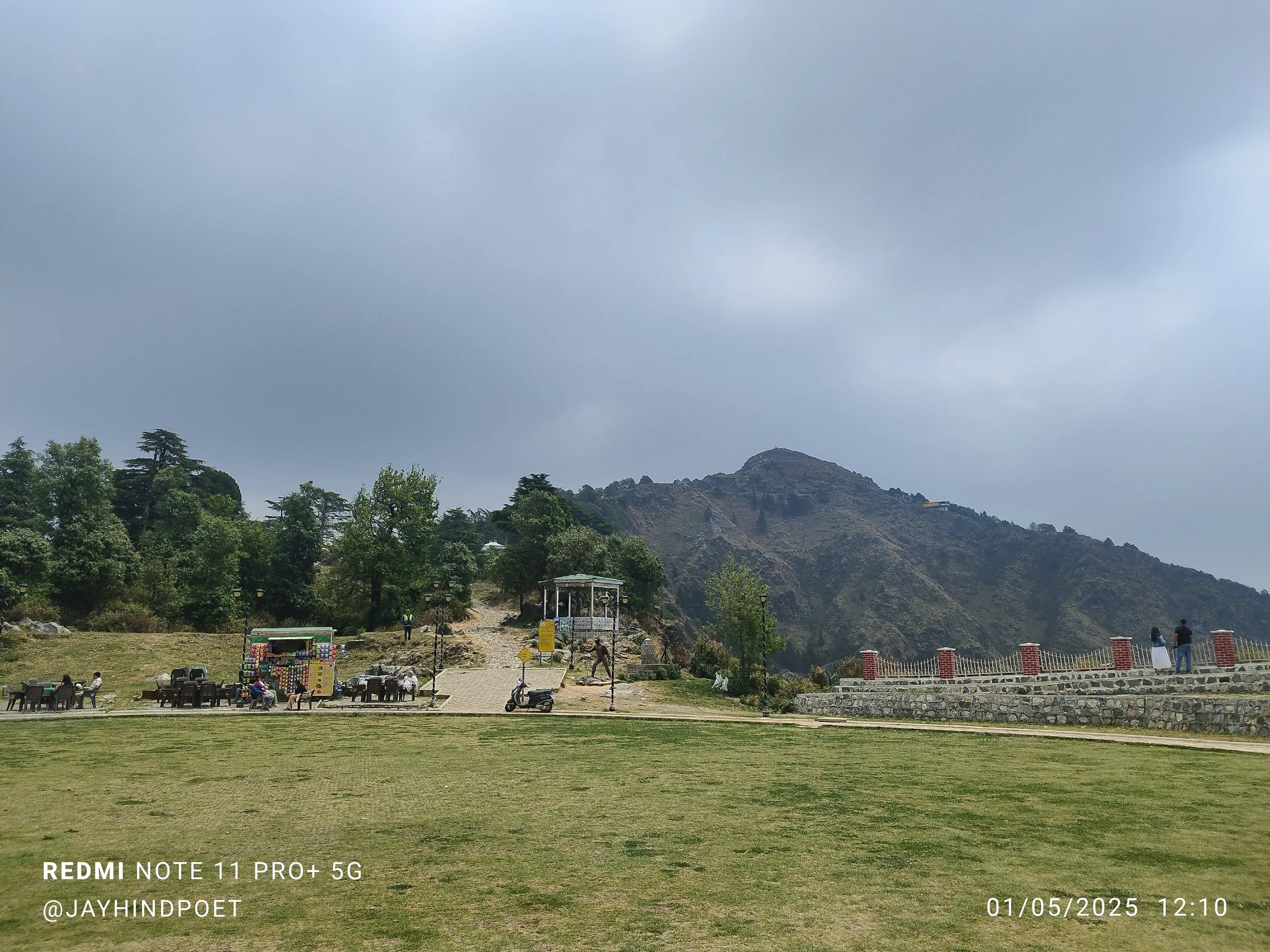 Lawn of the George Everest heritage site with food stalls and a gazebo, the stone path leading up toward the bare George Everest Peak under grey cloud