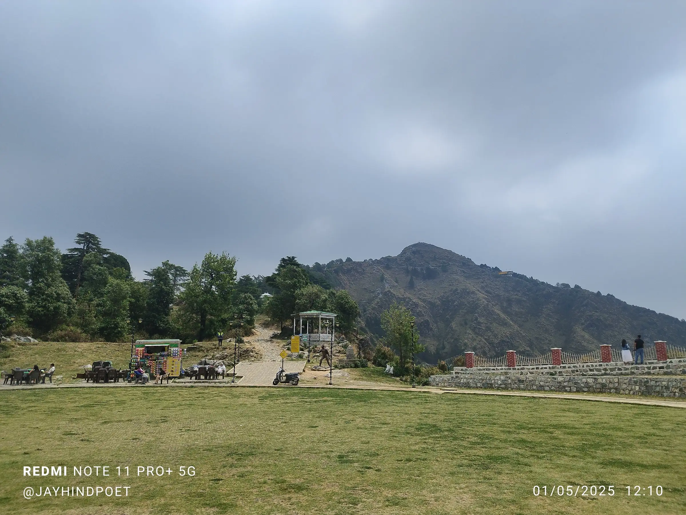 Lawn of the George Everest heritage site with food stalls and a gazebo, the stone path leading up toward the bare George Everest Peak under grey cloud