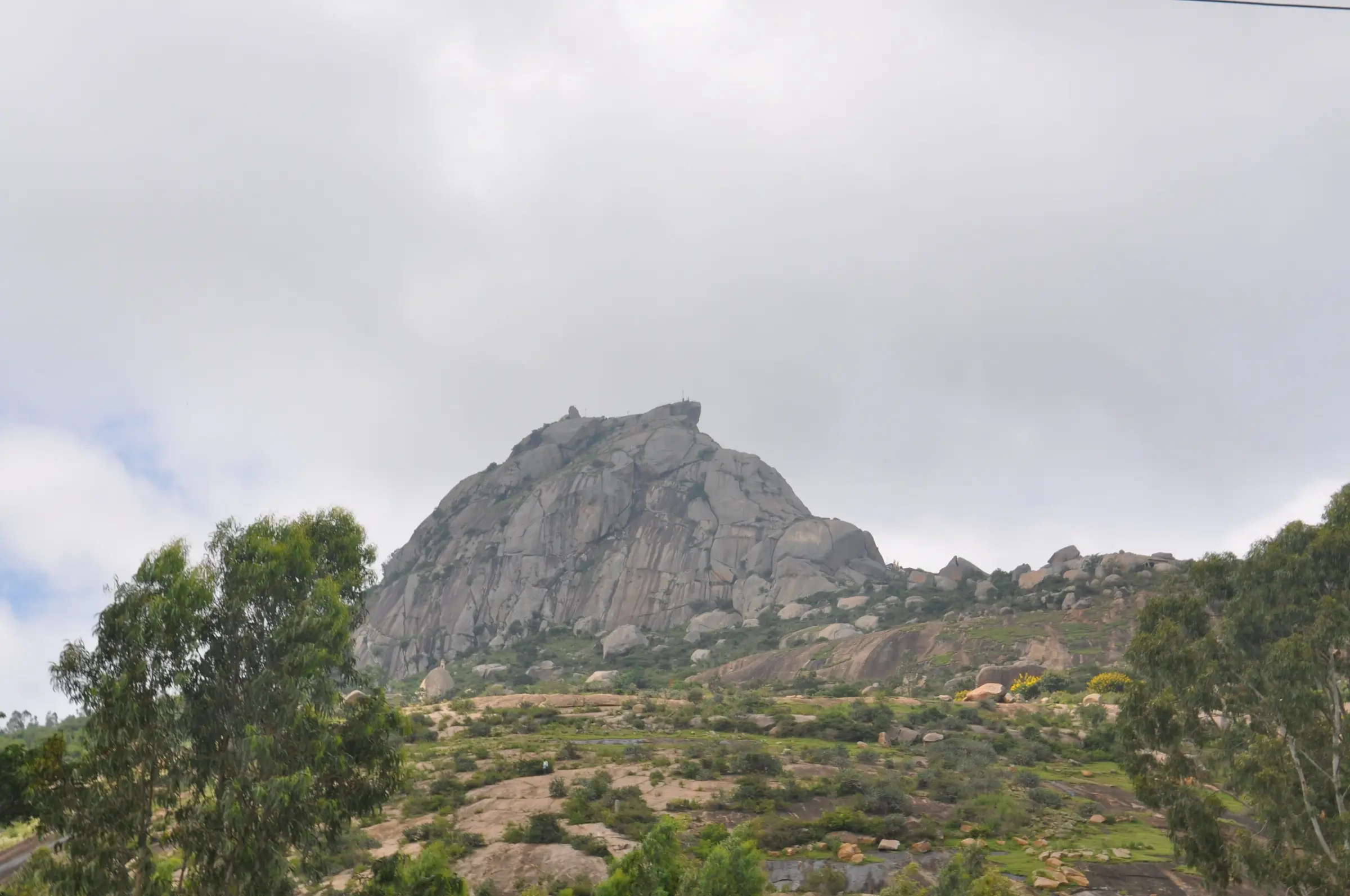 The bare granite dome of Shivagange hill rising above green scrub and eucalyptus trees under a grey sky, seen from the foothills