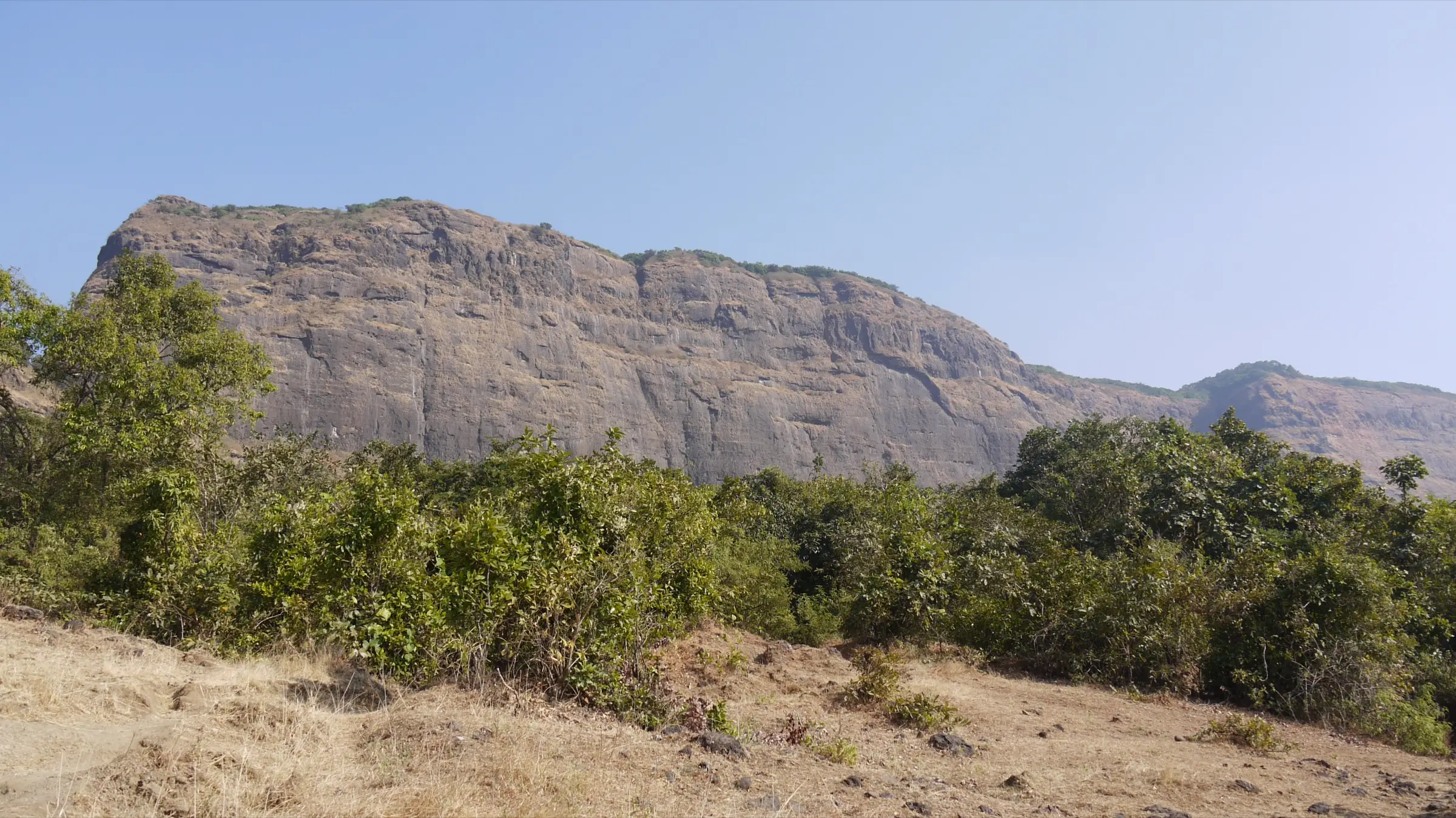 The long rock wall of Prabalgad fort rising above green scrub and dry grass, seen from the Prabalmachi plateau