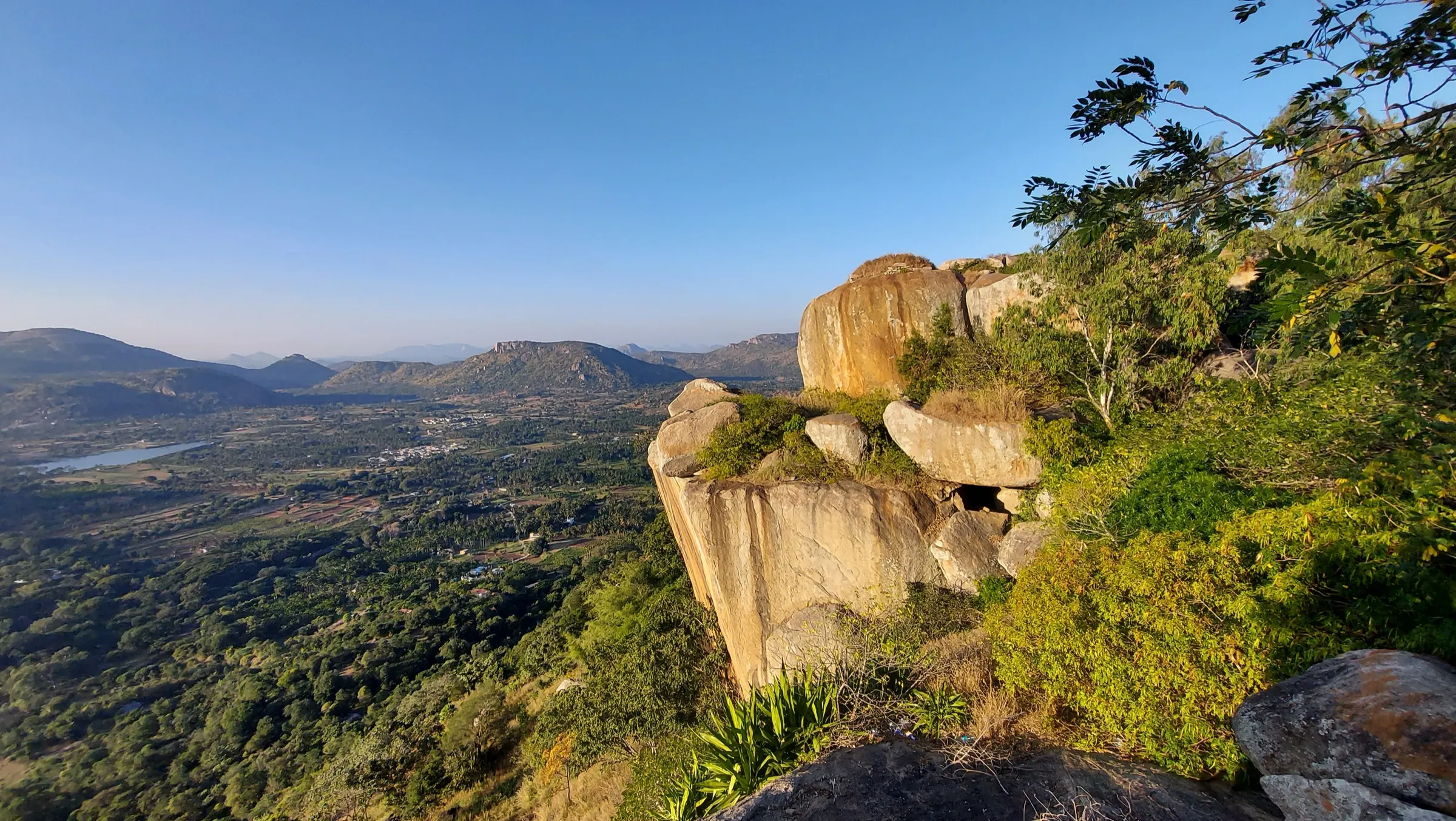 Granite boulders on the edge of Devarayanadurga hill in morning light, looking out over green farmland, a lake and rows of rocky hills