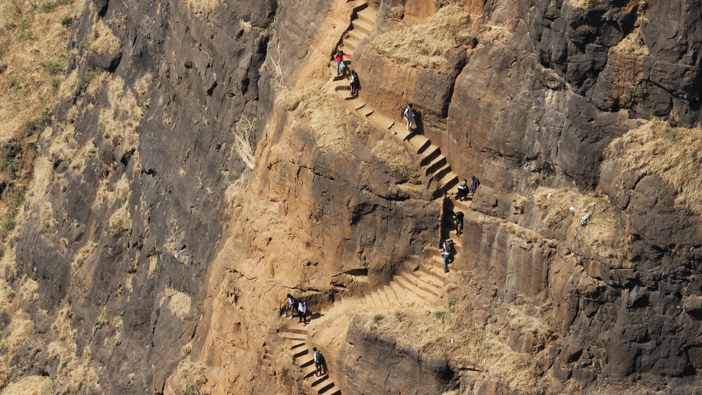 Trekkers climbing the narrow rock-cut steps that zig-zag up the face of the Kalavantin pinnacle