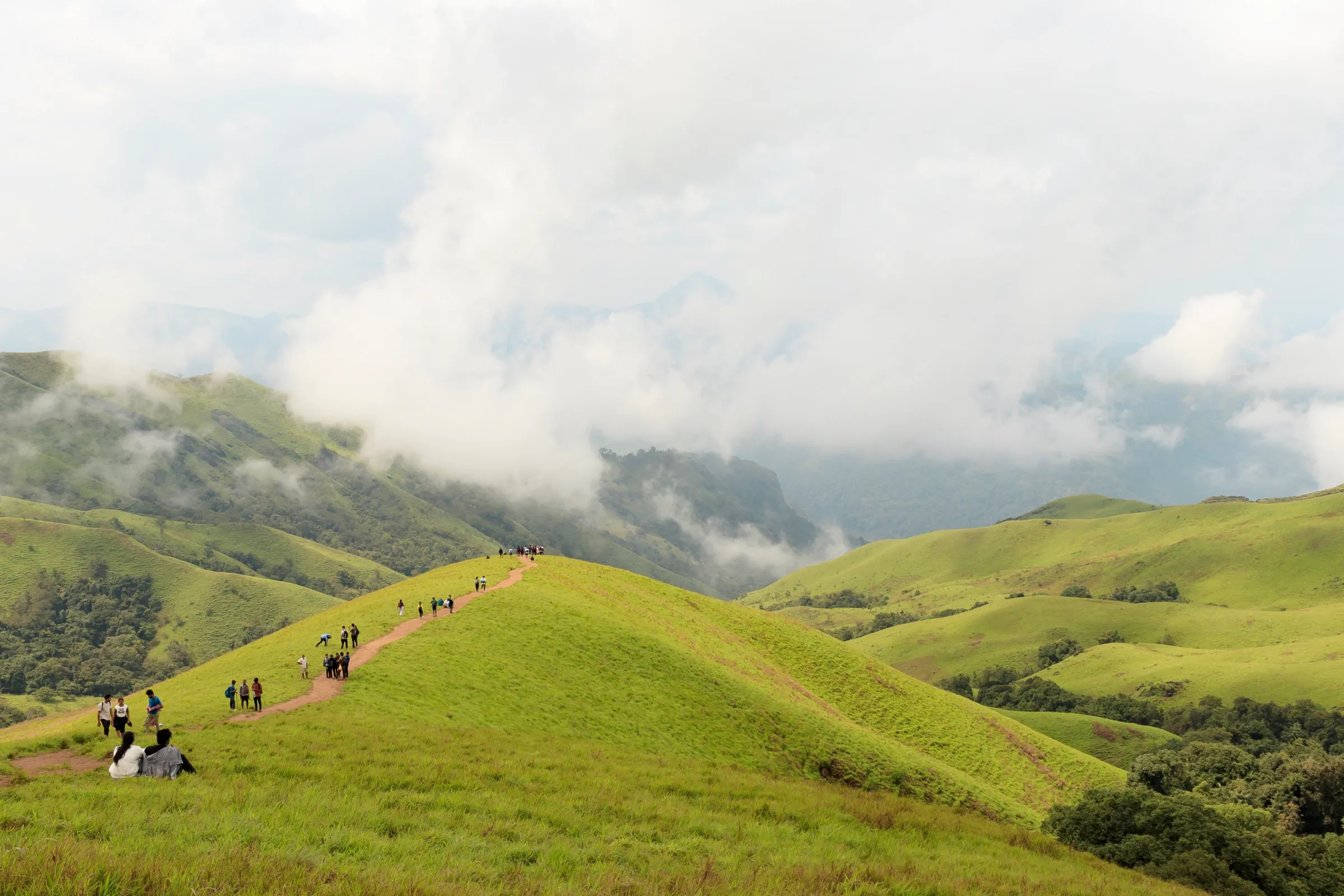 Trekkers walking a dirt trail along a green grassy ridge below the Netravathi summit, with clouds over the hills of the Kudremukh range