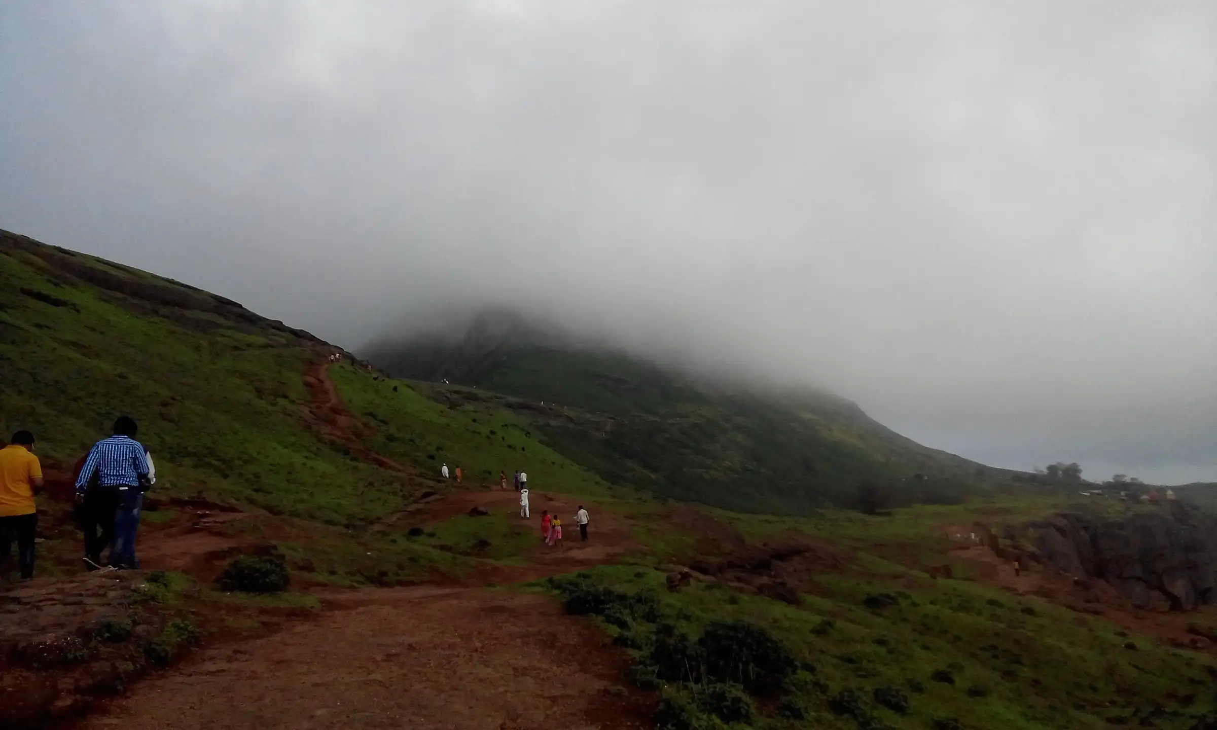 Walkers on a red-earth path across green grassy slopes of Brahmagiri, with the upper hill hidden in monsoon cloud