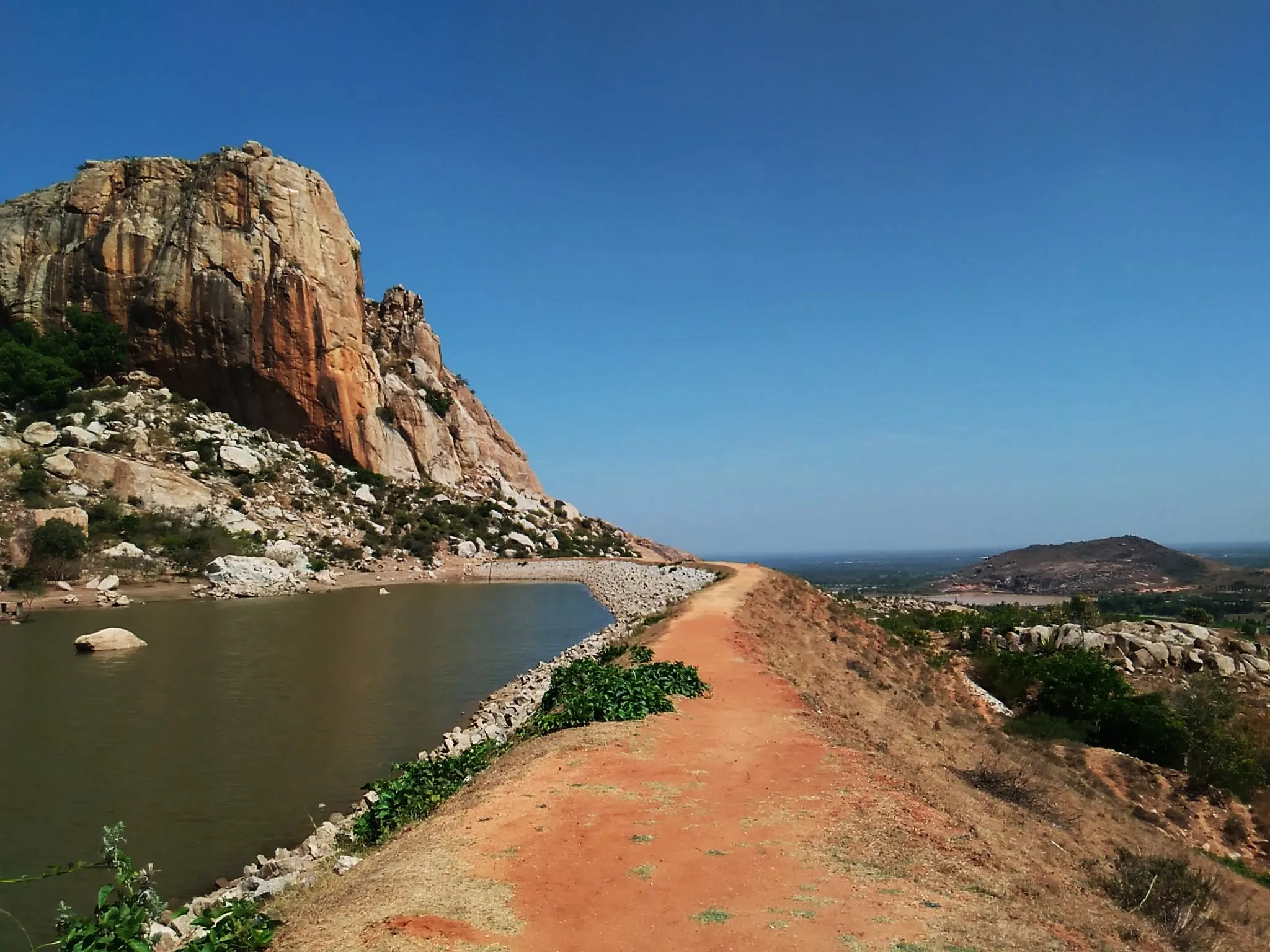 Red-earth path along a tank bund below the granite cliffs of Kailasagiri, the hill next to Ambaji Durga, with the Chintamani plain beyond (nearby area, not the trek itself)