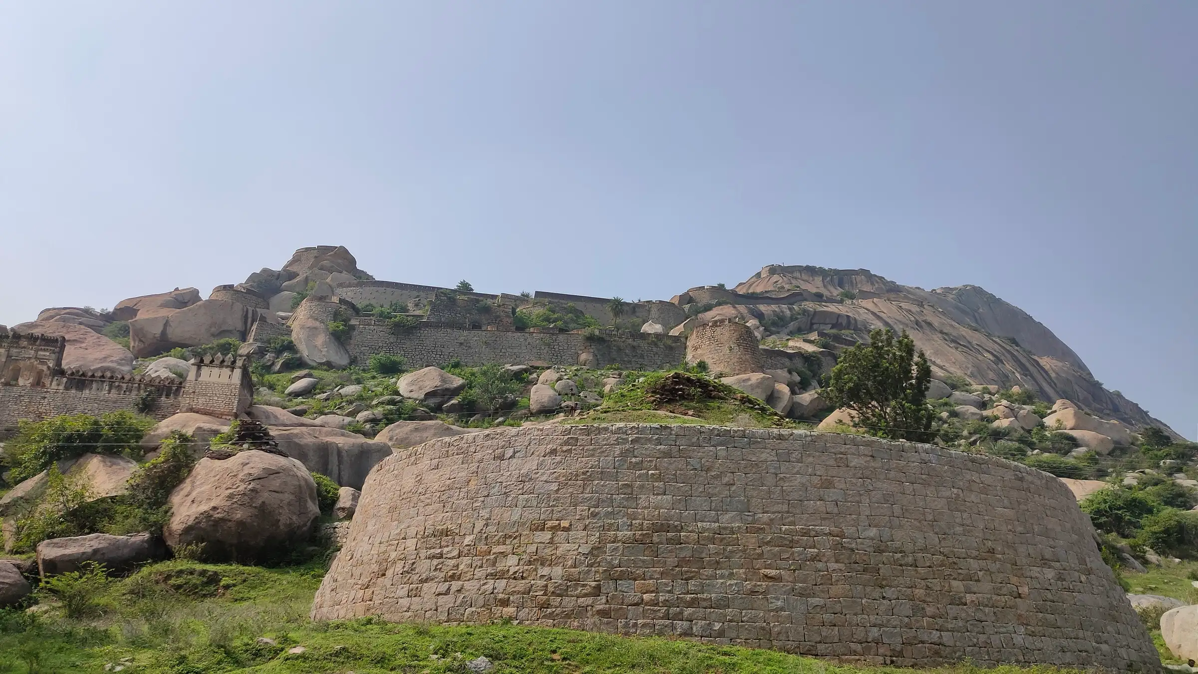 Round stone bastion and fort walls on the lower slopes of the Madhugiri granite monolith, with the bare rock dome rising behind