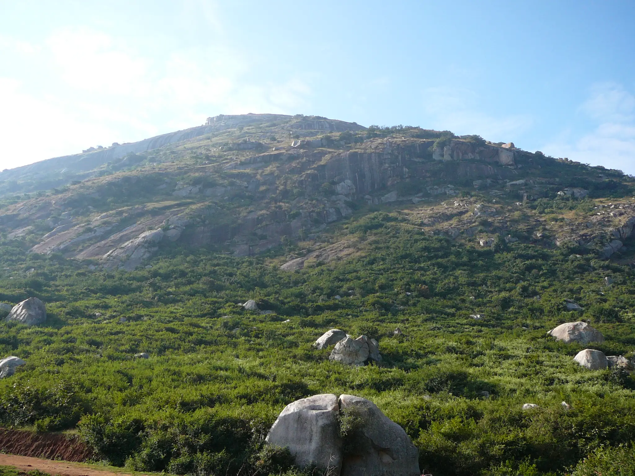 Skandagiri hill seen from the base, rock faces above green shrubland dotted with boulders