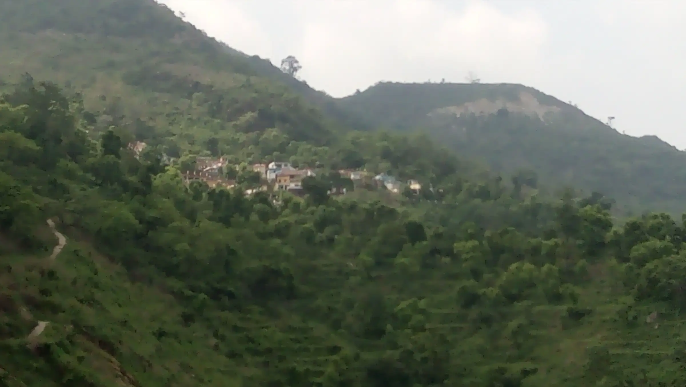 A narrow footpath winding across a green, forested hillside below a small village near Neelkanth