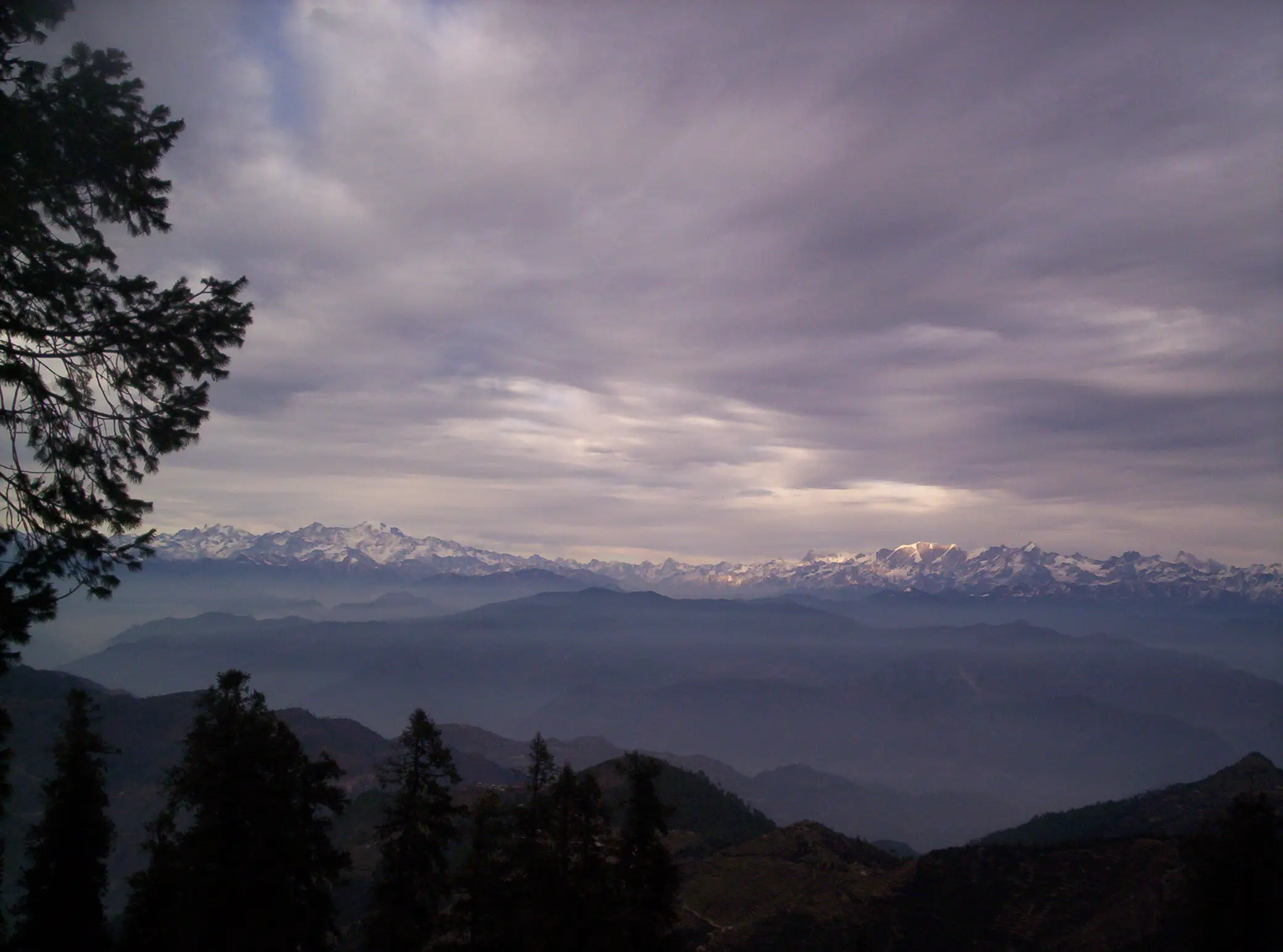 Snow-capped Himalayan range under grey cloud seen beyond dark forested ridges from Surkanda Devi