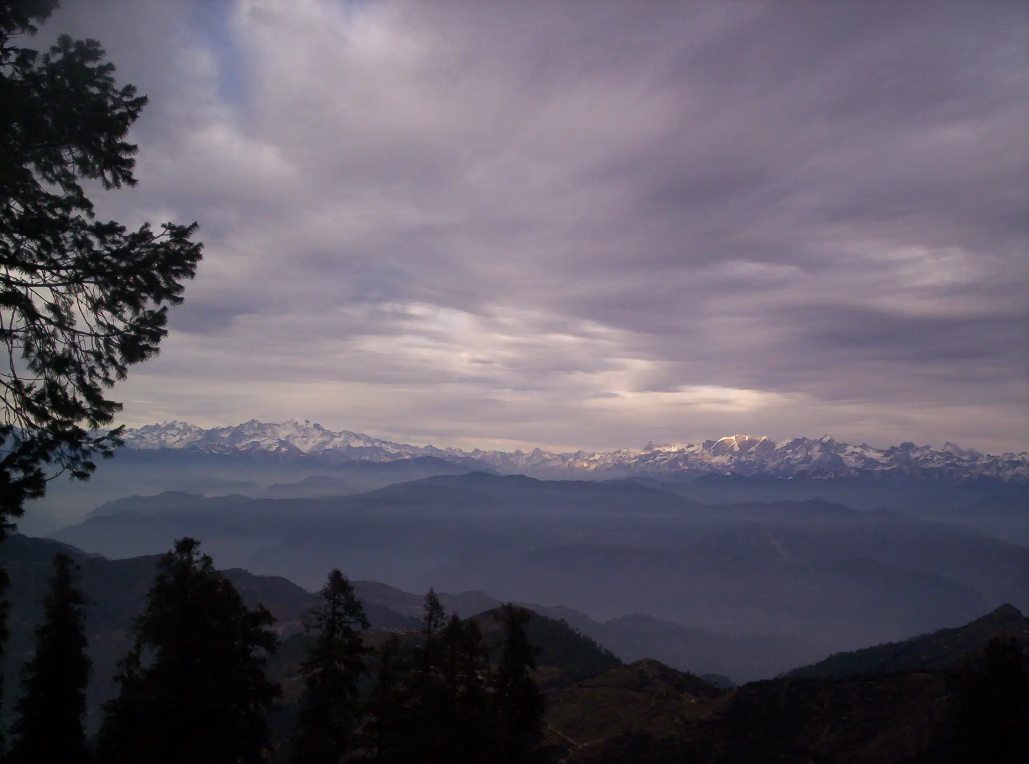 Snow-capped Himalayan range under grey cloud seen beyond dark forested ridges from Surkanda Devi