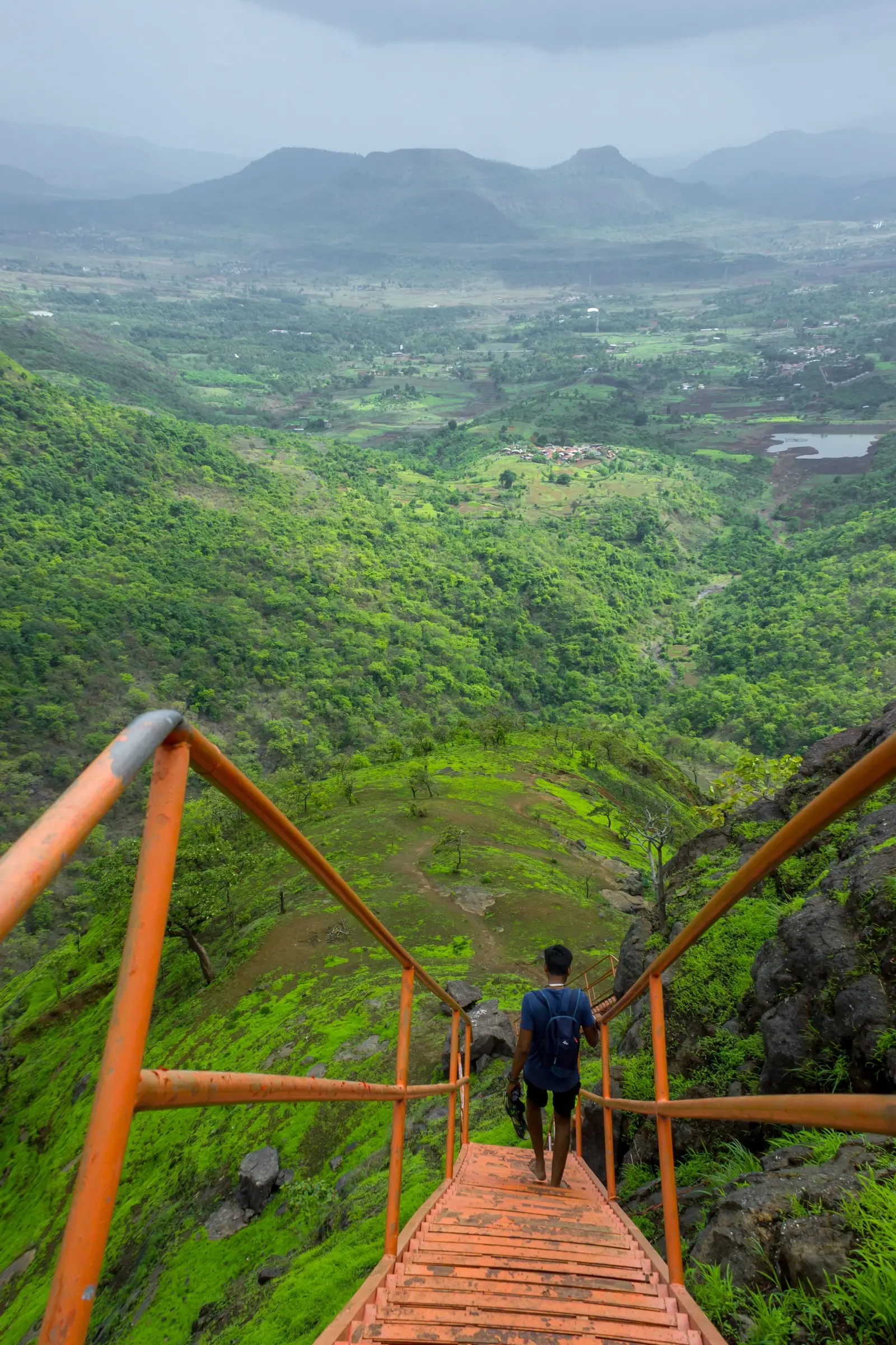 A trekker walking down the orange iron ladder of Sondai Fort towards green hills, villages and a small reservoir