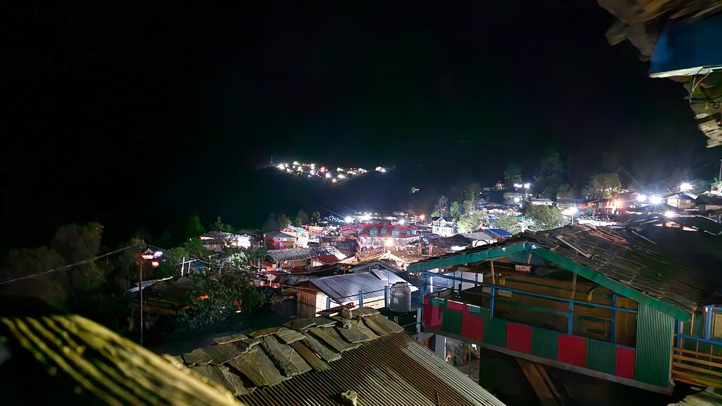 Jakhol village at night with lit houses and slate roofs, and a distant village glowing on the hillside
