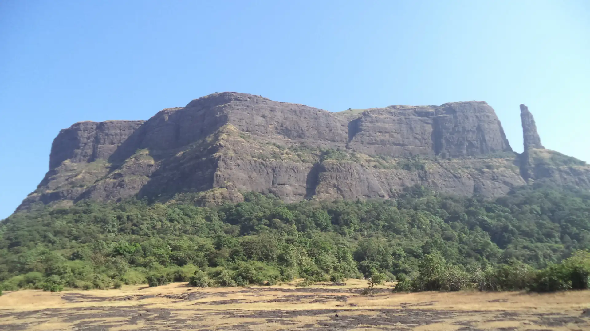 The flat-topped rock wall of Jivdhan Fort with the Vanarlingi pinnacle at its right end, seen across a dry plateau from Naneghat