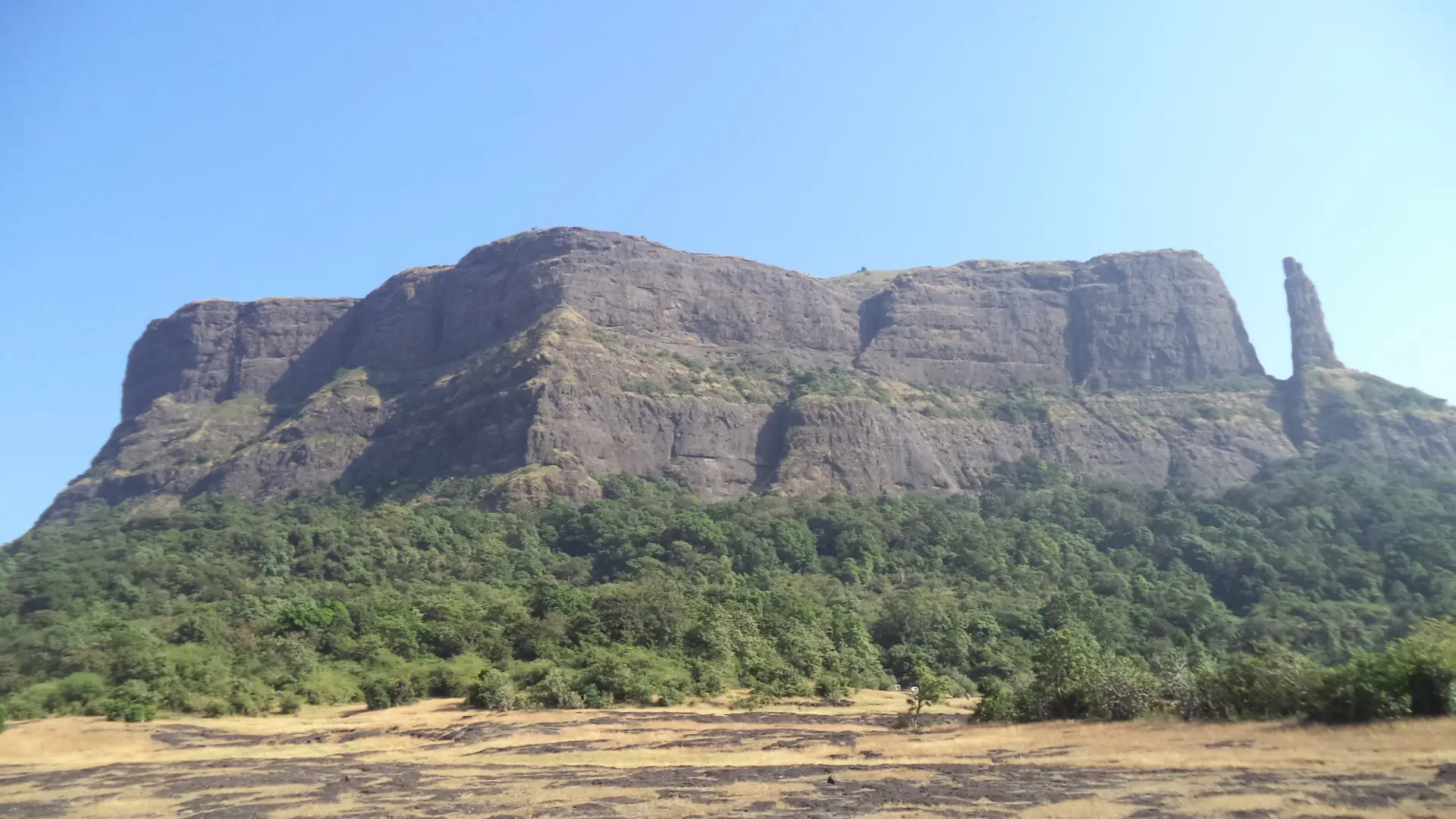 The flat-topped rock wall of Jivdhan Fort with the Vanarlingi pinnacle at its right end, seen across a dry plateau from Naneghat