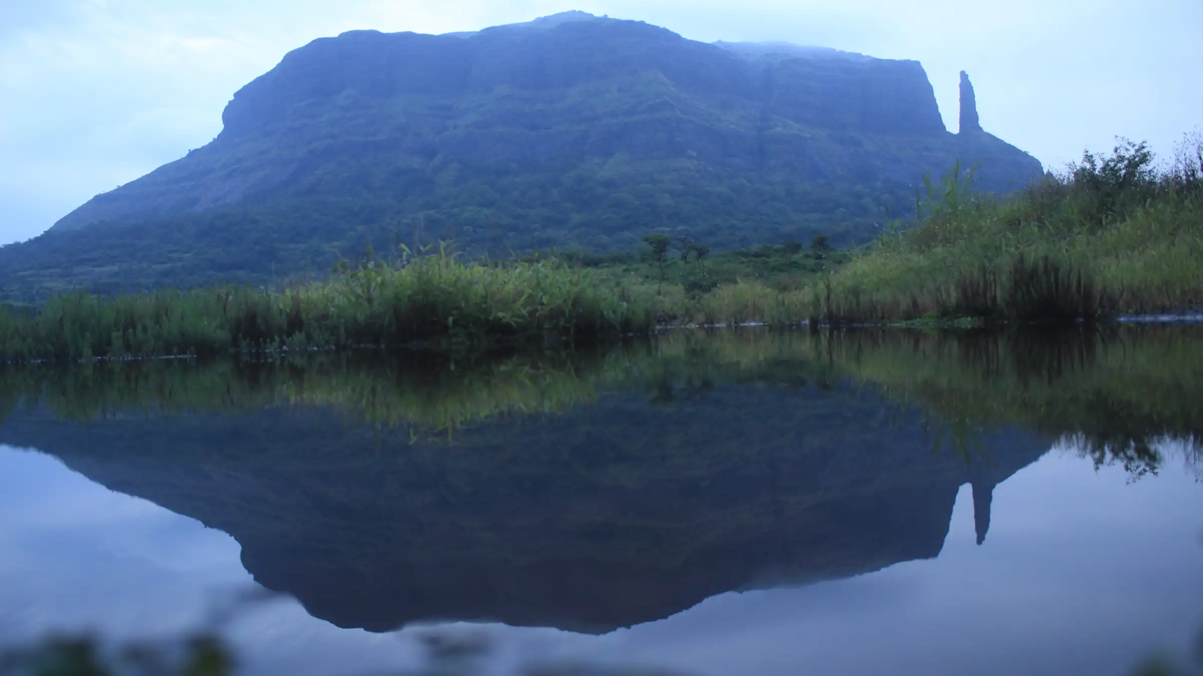 Jivdhan hill and the Vanarlingi pinnacle reflected in a still pond under a cloudy sky