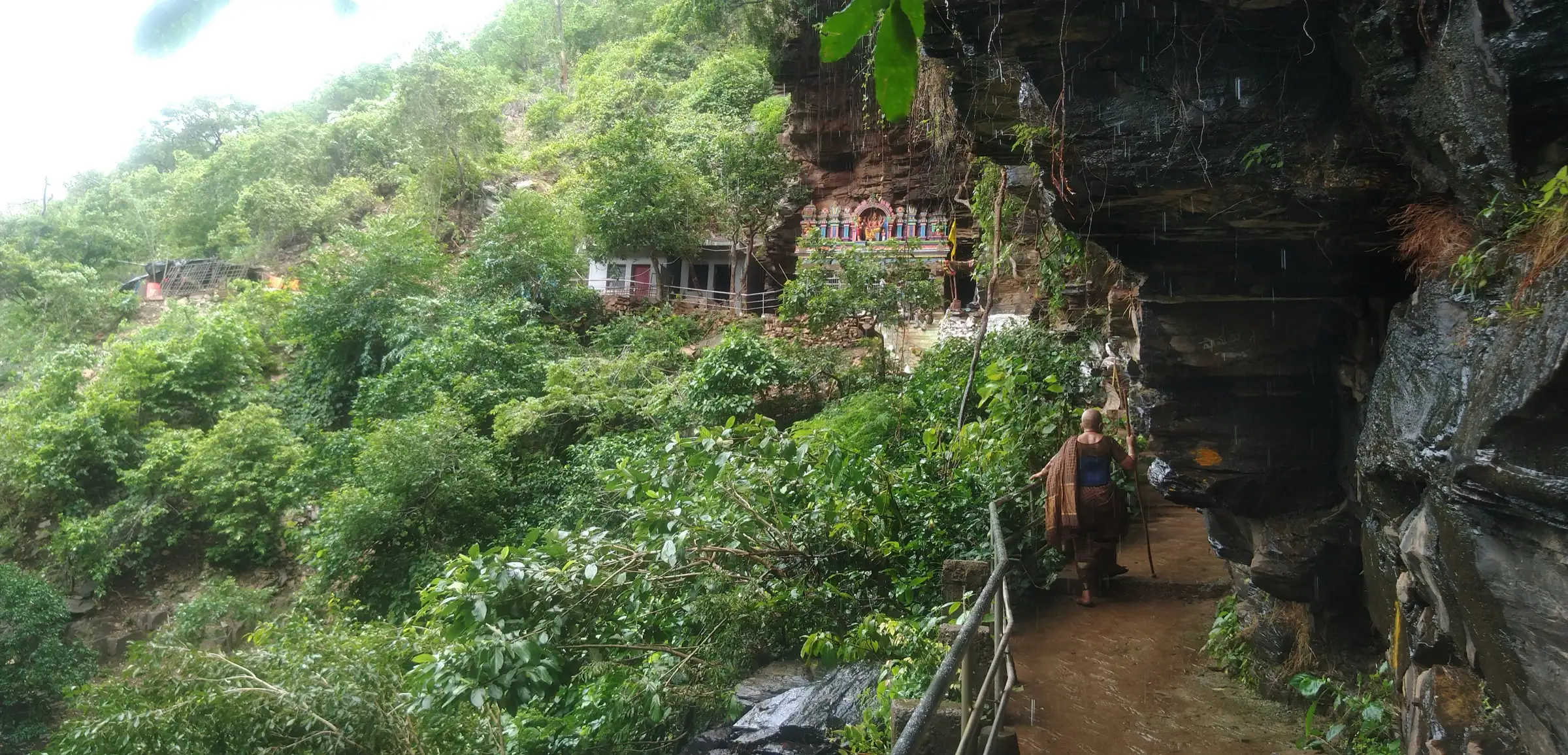 Railed rock path under an overhanging cliff leading to Jwala Narasimha temple on a forested hillside