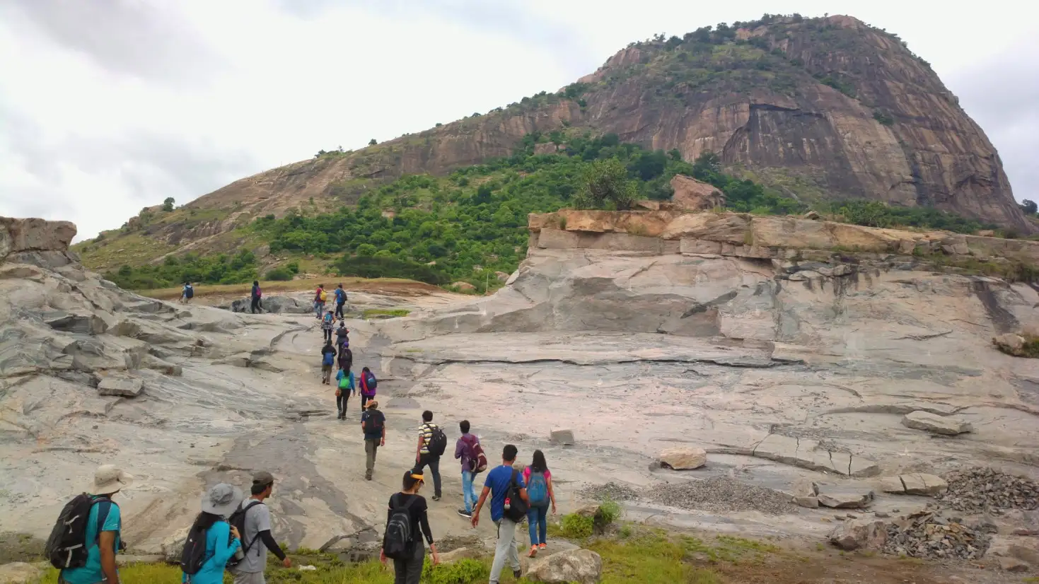 Trekkers crossing bare granite slabs at the start of the hike, with the Kabbaladurga monolith rising behind under a cloudy sky