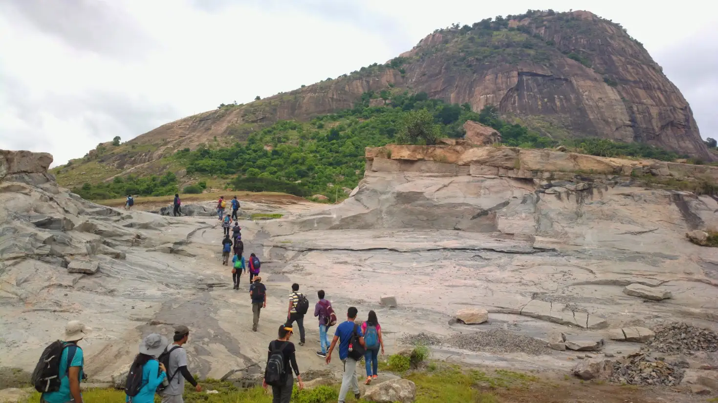 Trekkers crossing bare granite slabs at the start of the hike, with the Kabbaladurga monolith rising behind under a cloudy sky