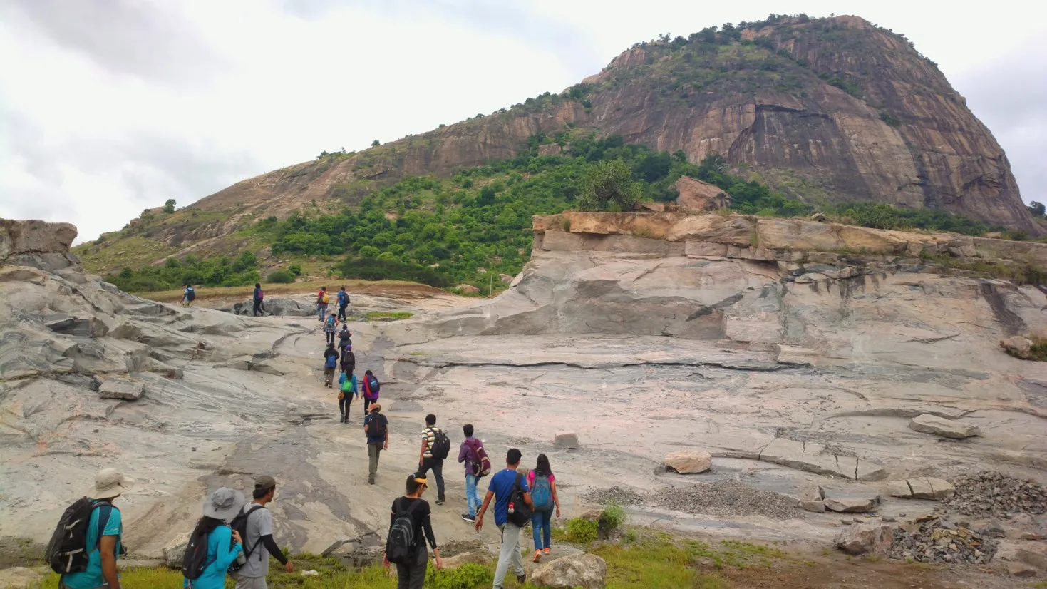 Trekkers crossing bare granite slabs at the start of the hike, with the Kabbaladurga monolith rising behind under a cloudy sky