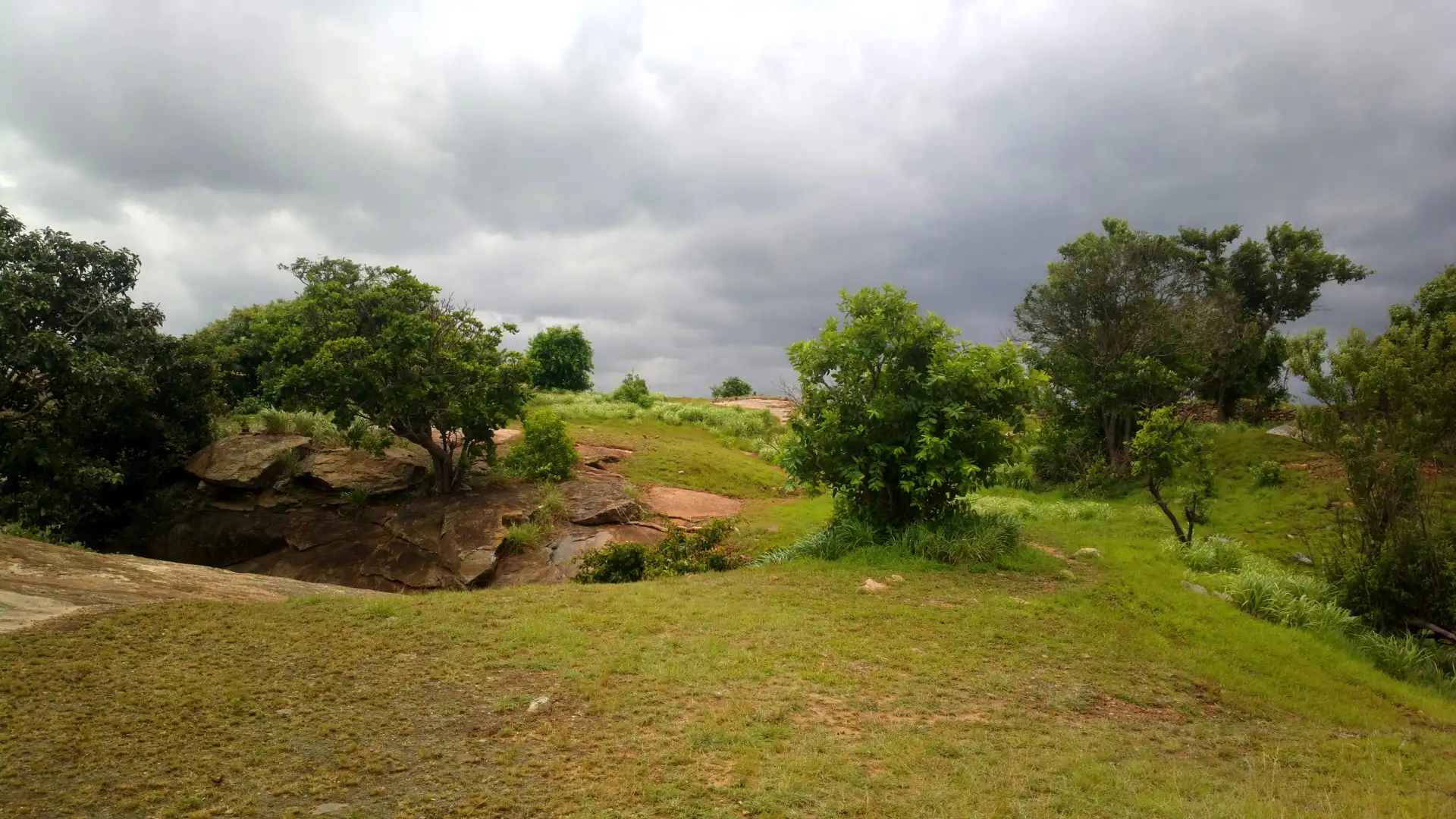 Grassy, tree-dotted flat top of Kabbaladurga with exposed granite under monsoon clouds