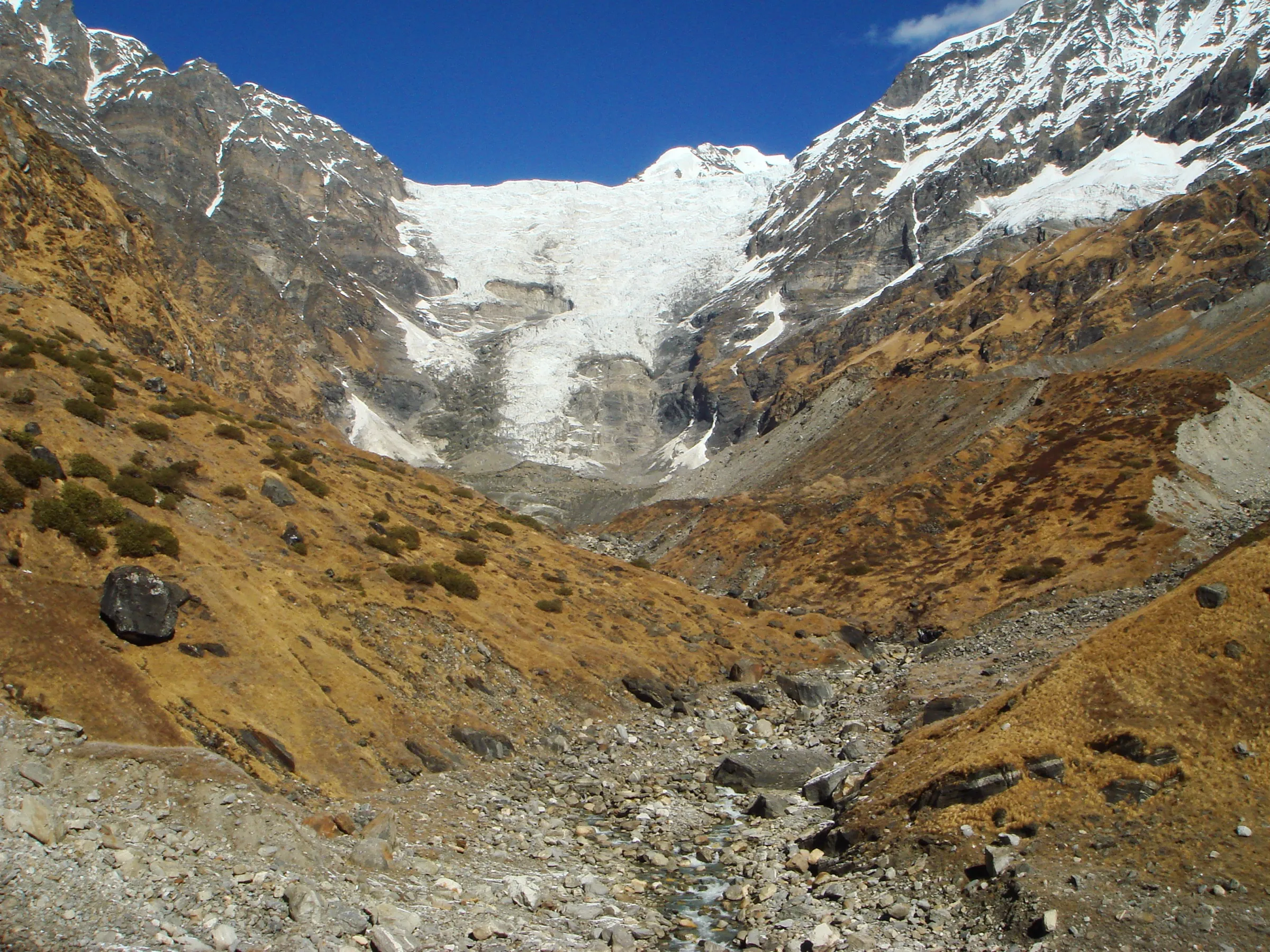 The white icefall of the Kafni Glacier pouring down between rock walls under a blue sky, above a boulder-strewn stream bed and brown autumn grass slopes