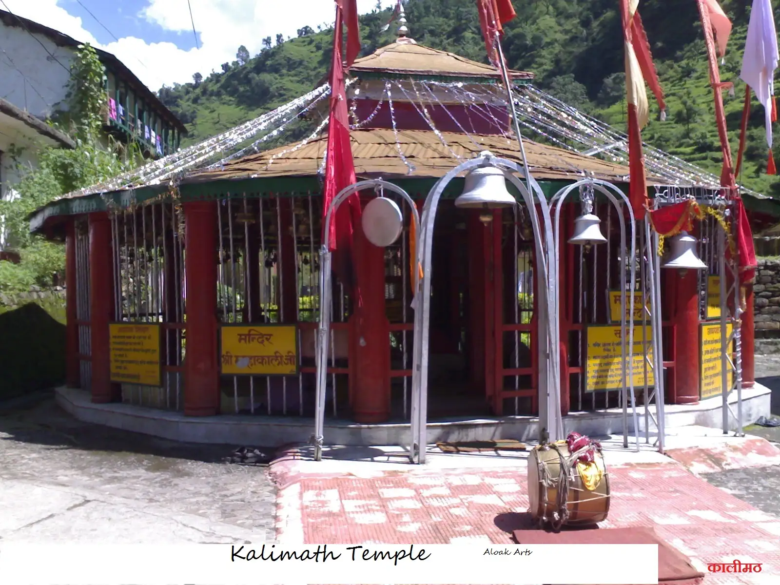 The red-pillared Maa Mahakali mandir at Kalimath with brass bells and flags, below green terraced hills