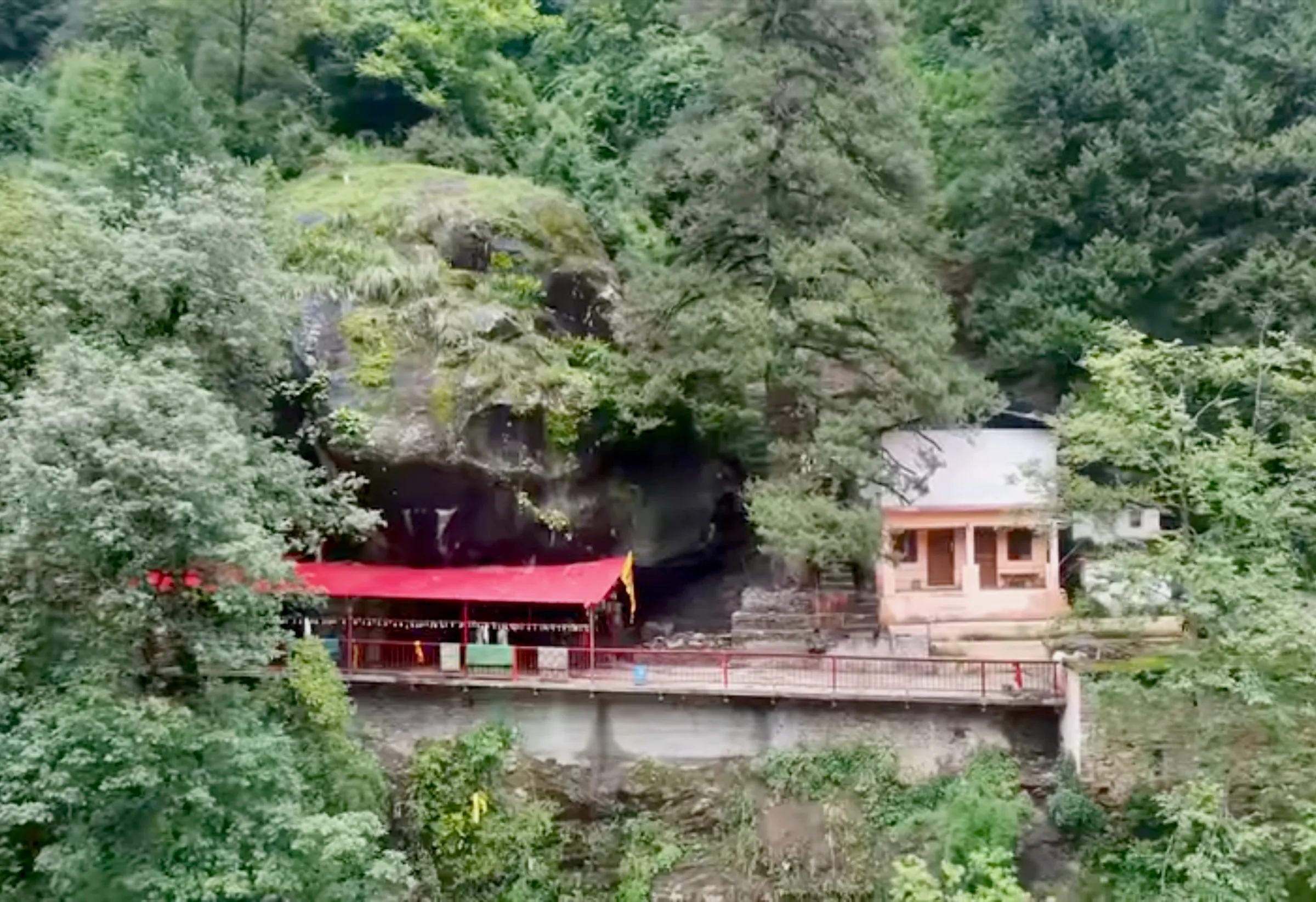 Kalpeshwar cave temple below a large mossy rock overhang, with a red-roofed shelter and railing among dense green forest