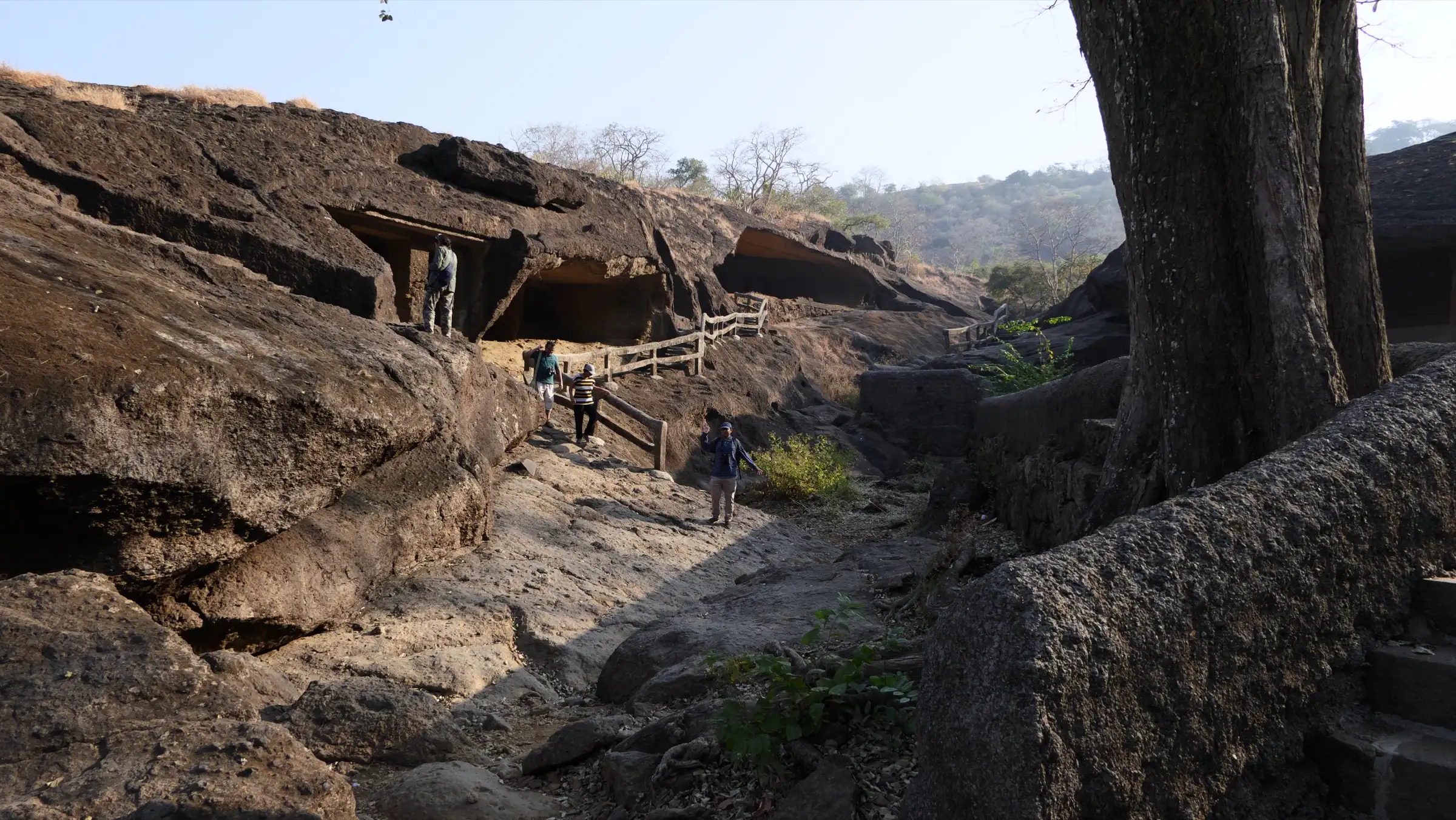 Walkers on a rocky path with wooden railings between rock-cut cave openings on the Kanheri hillside