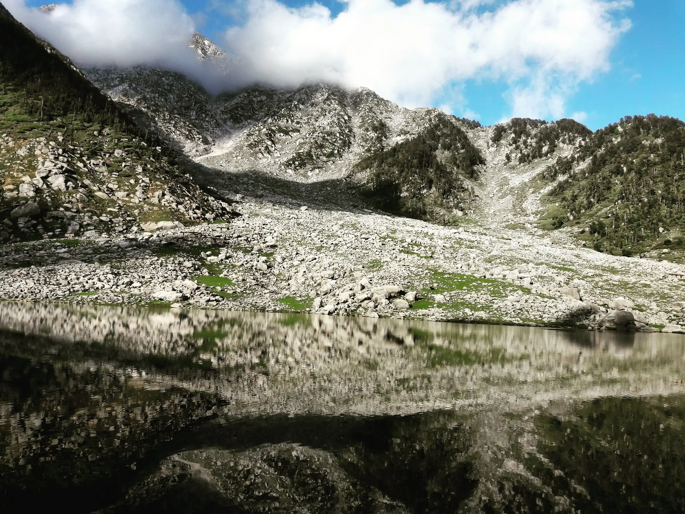Kareri Lake reflecting the boulder slopes and cloud-capped Dhauladhar ridge that rise towards Minkiani Pass