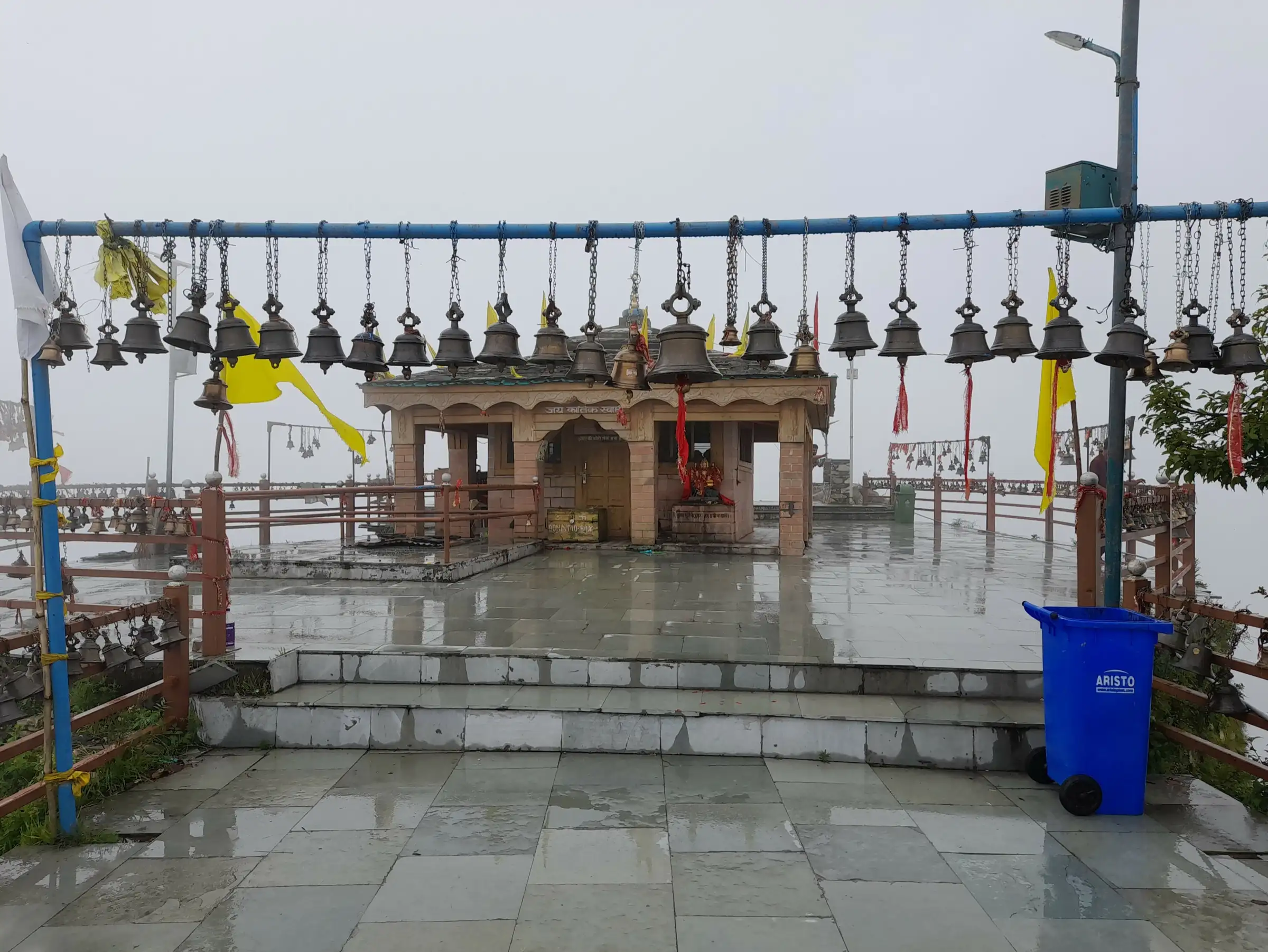 Kartik Swami temple courtyard on a misty day, with rows of bells hanging from a railing in front of the stone shrine