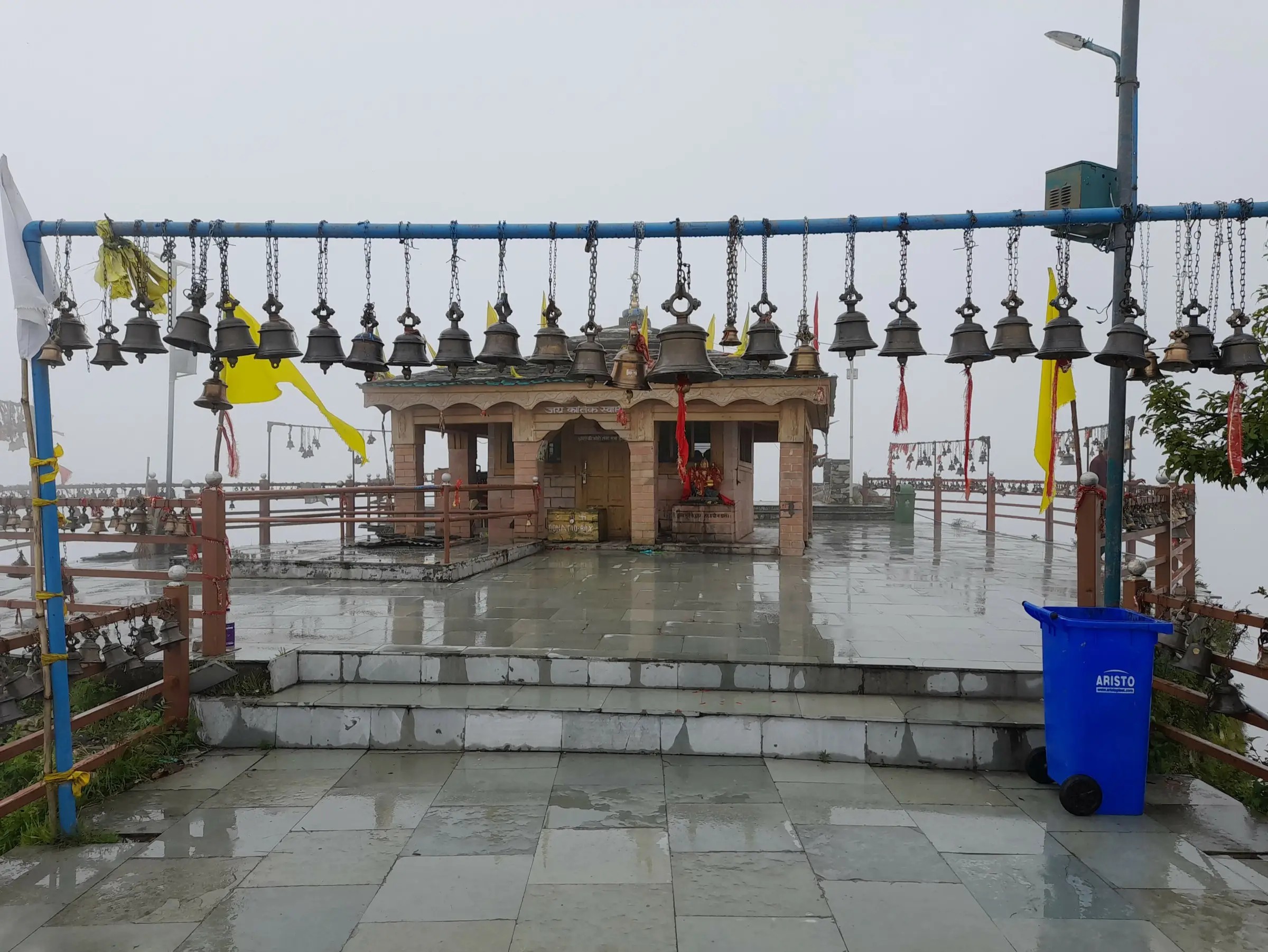 Kartik Swami temple courtyard on a misty day, with rows of bells hanging from a railing in front of the stone shrine