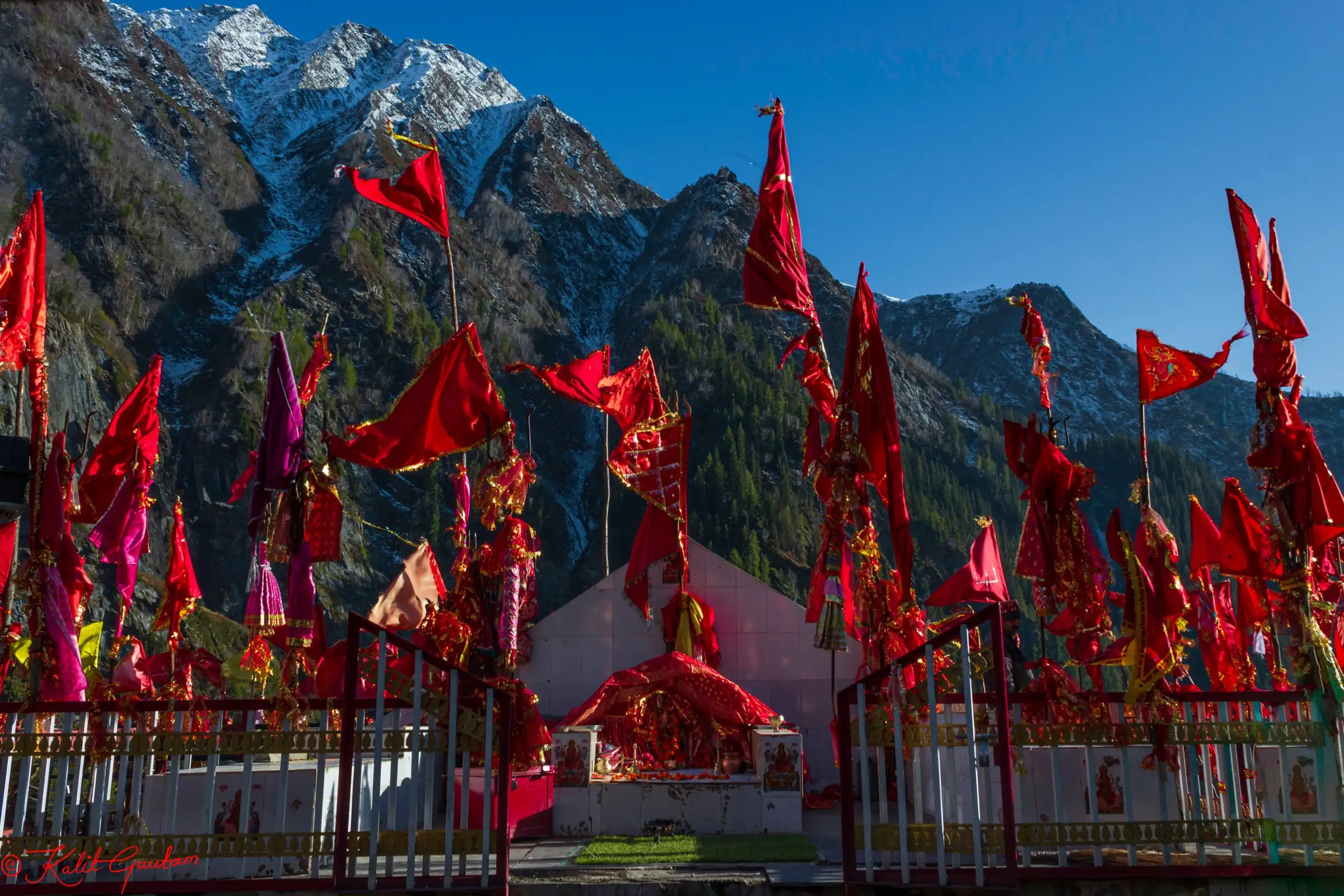 A Devi shrine hung with red flags, photographed during the Kartik Swami yatra at Kugti, with snow-dusted peaks and pine slopes of the Budhil valley behind
