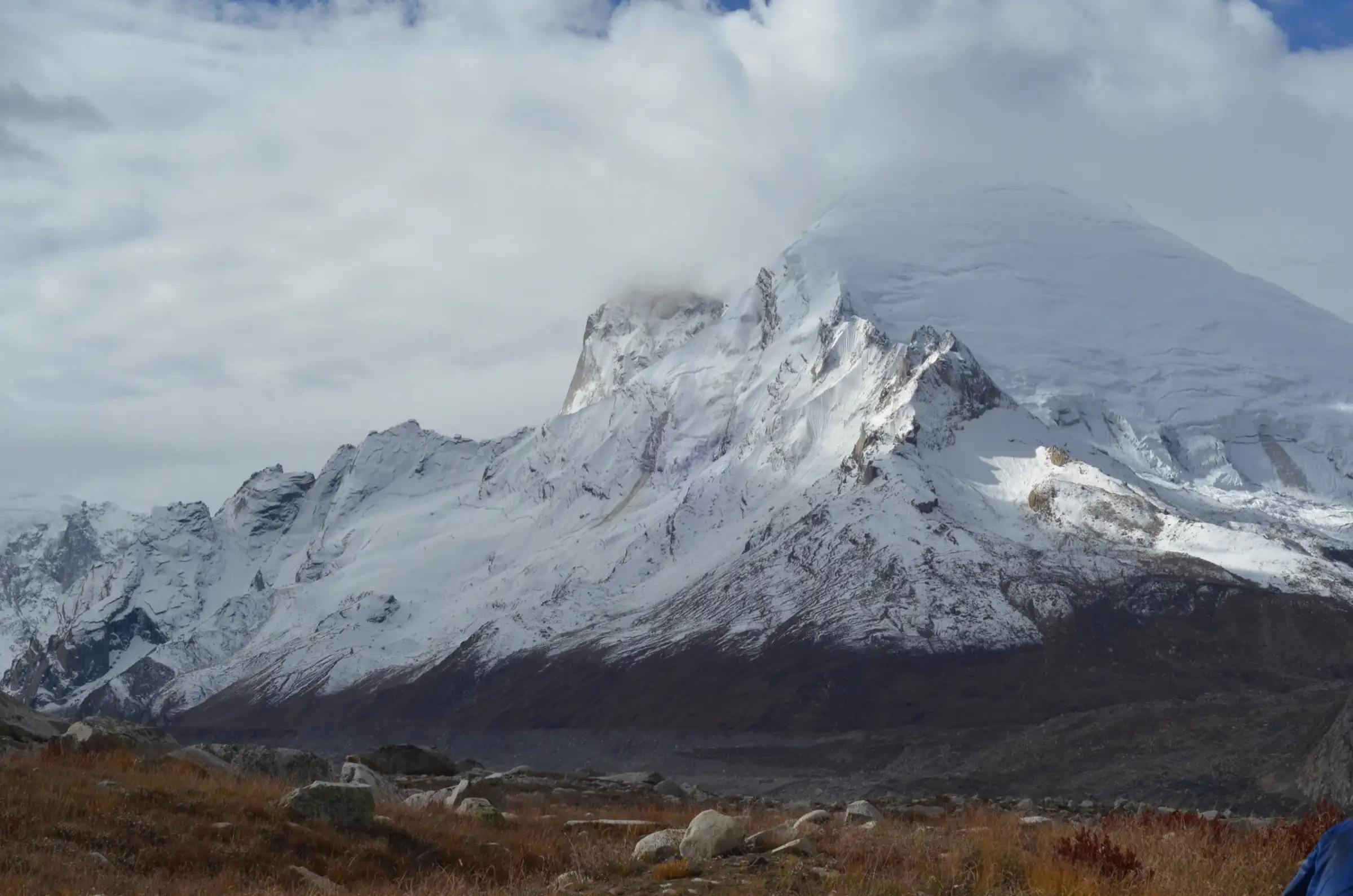 Snow-covered Kedar Dome partly in cloud, seen across autumn grass and boulders from Nandanvan
