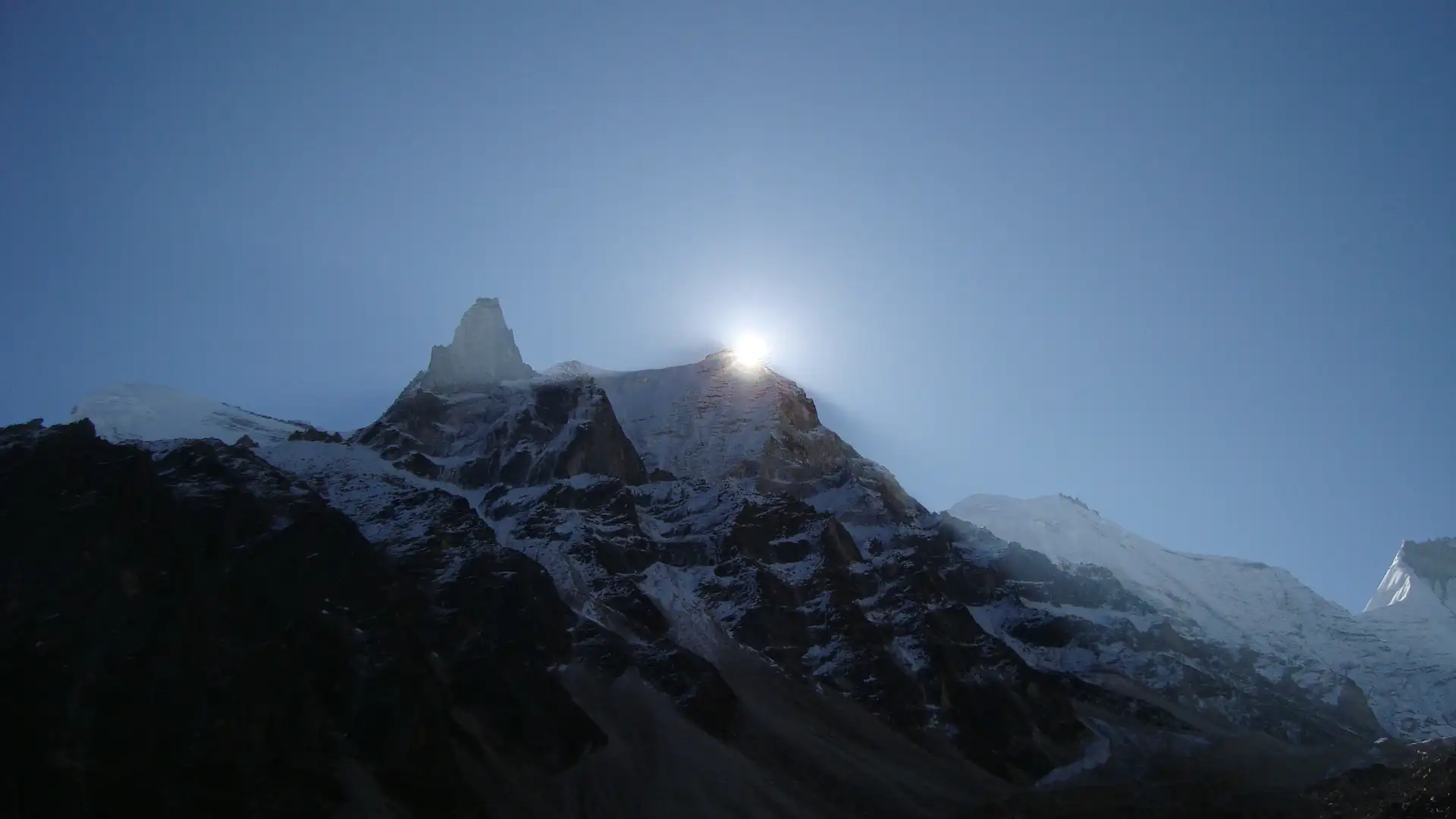 The sun rising behind a rocky snow-streaked peak seen from the Kedar Kharak camp