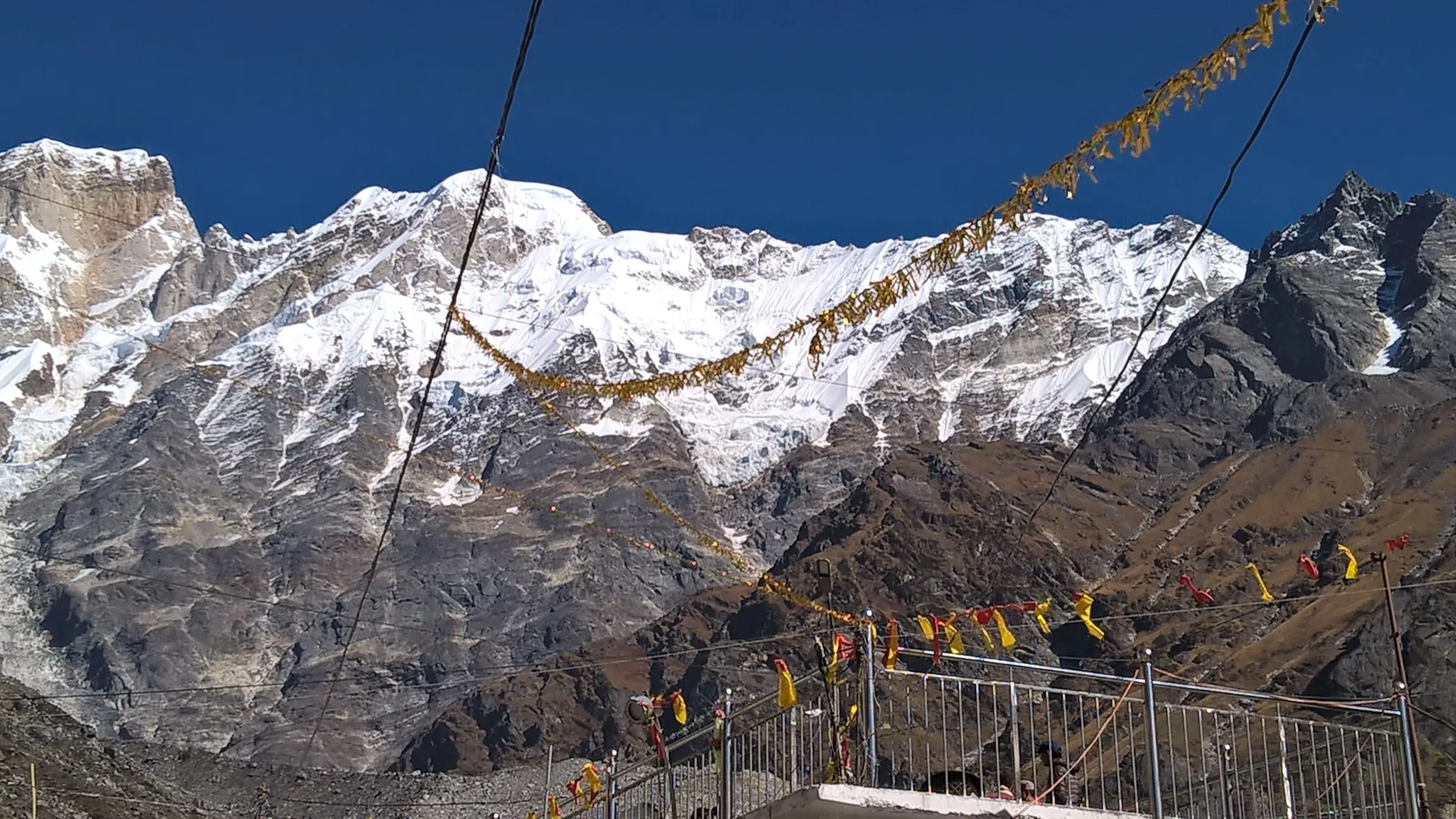 Snow-covered Kedarnath peak under a deep blue sky, with prayer garlands strung above a railing at Kedarnath