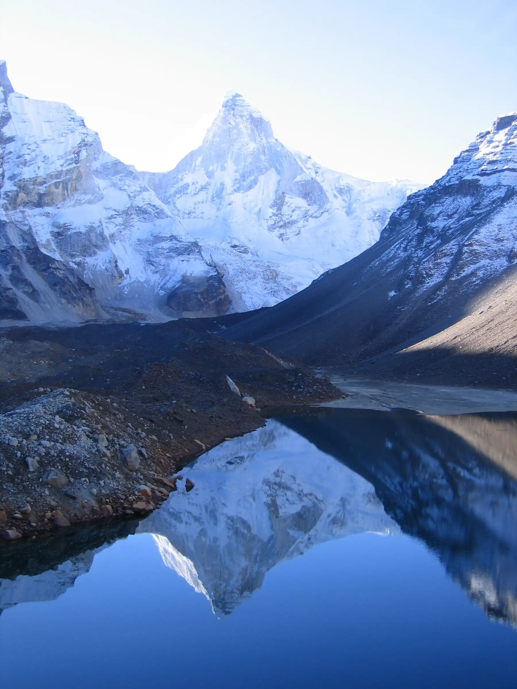 Thalay Sagar mirrored in calm blue water at Kedartal, with a dark moraine bank in the foreground
