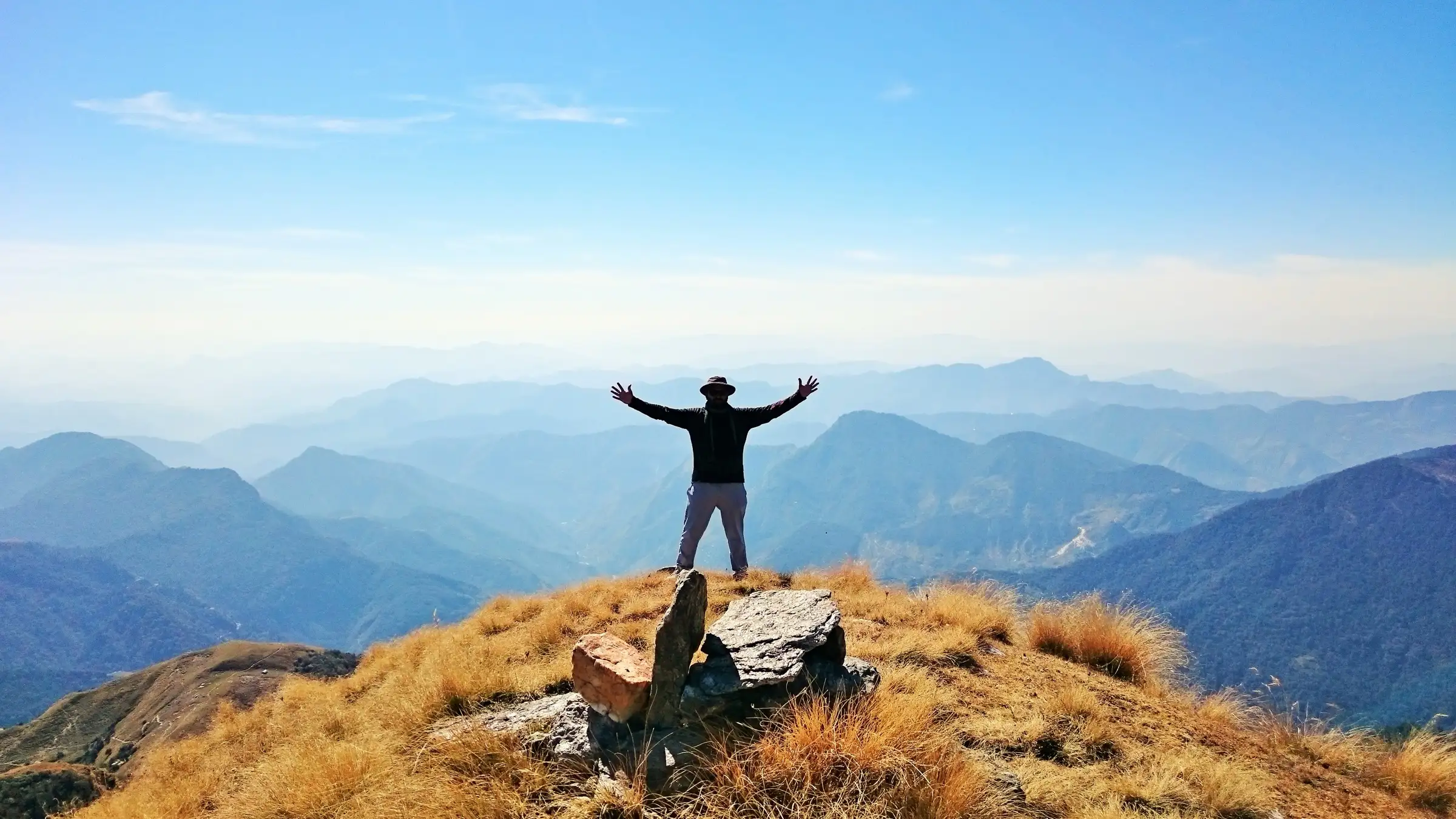 Trekker with arms raised on the grassy, golden top of Khalia peak above layers of blue Kumaon ridges