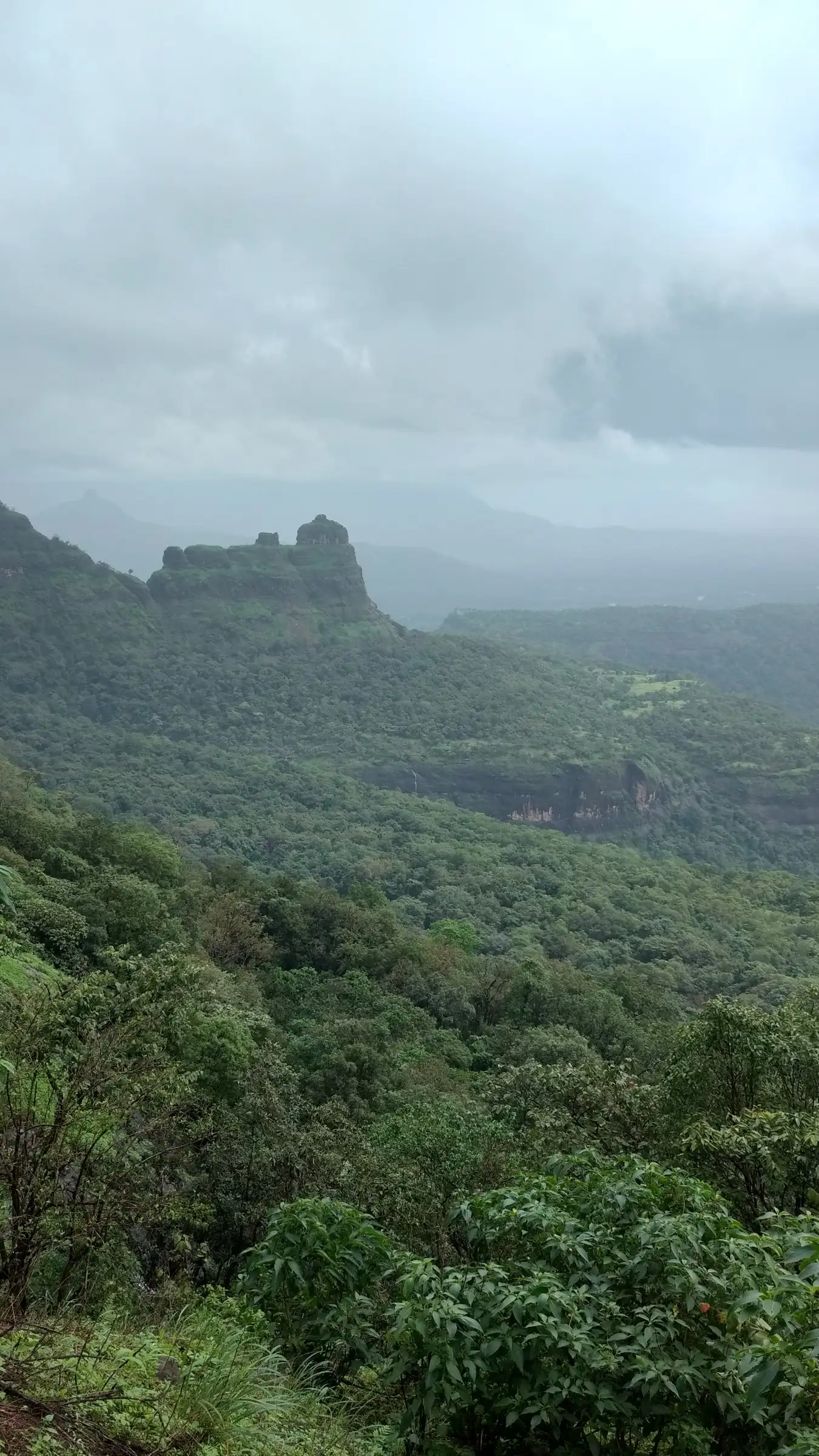 Misty monsoon view of forested slopes and a rock pinnacle seen from the trail above Khandas