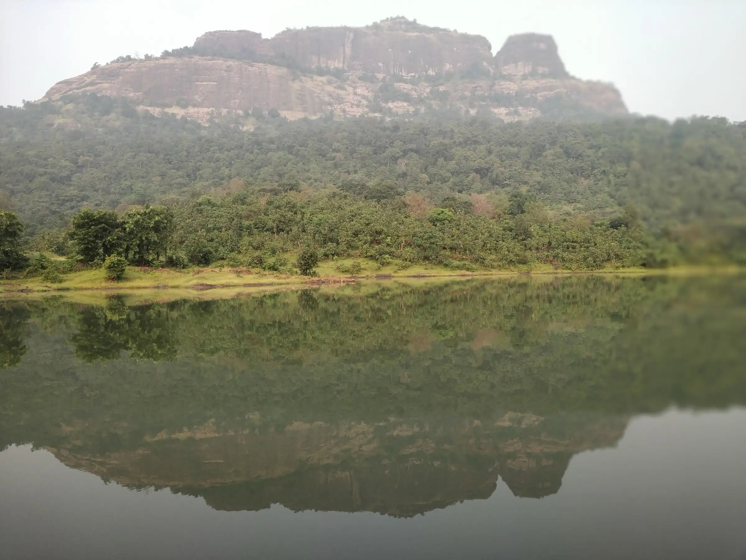 The rock cliffs of Kohoj Fort above a forested slope, reflected in the still lake at its foot