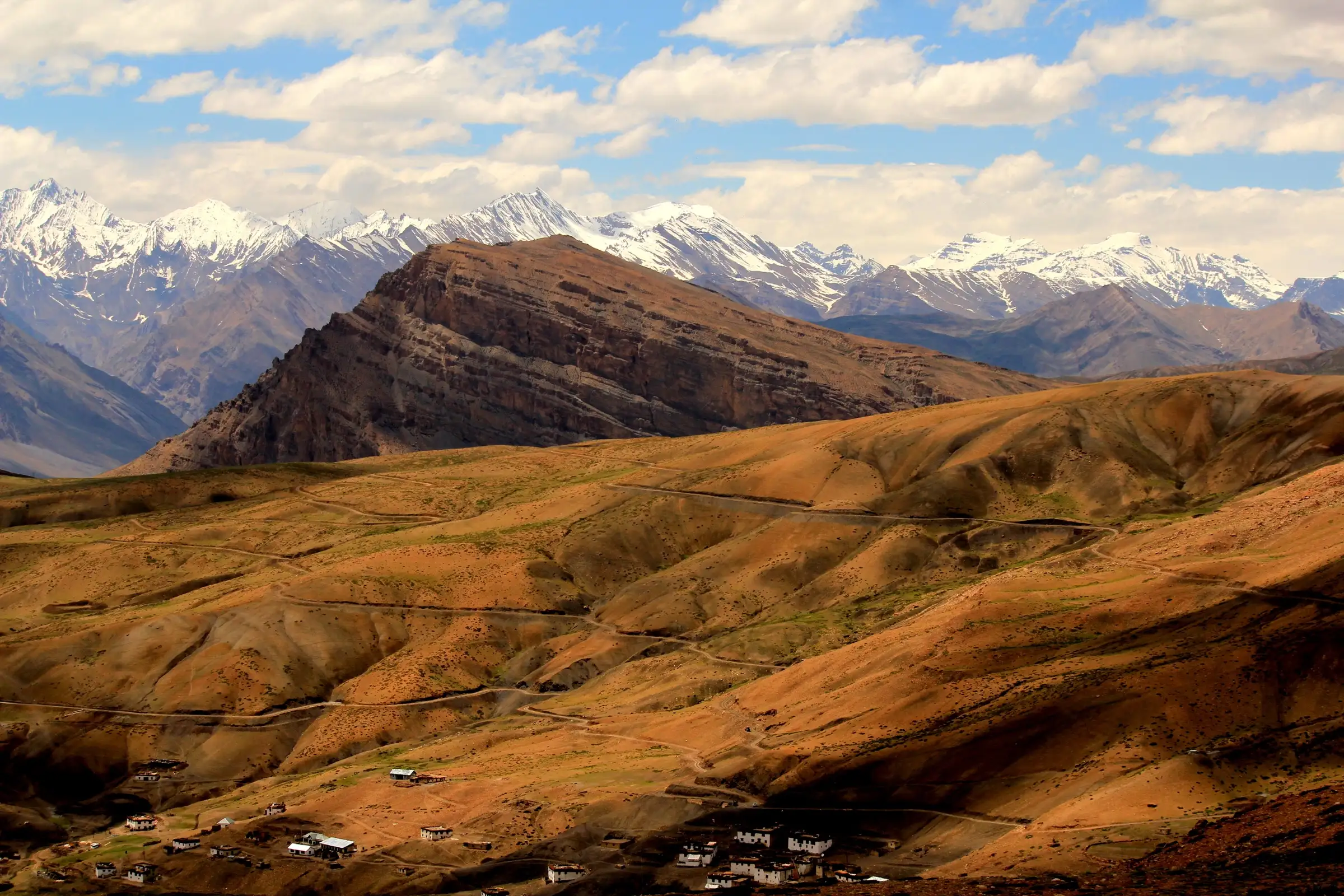 Brown, terraced slopes with a winding dirt road above the houses of Komic village, with a banded rock ridge and snow peaks of Spiti behind