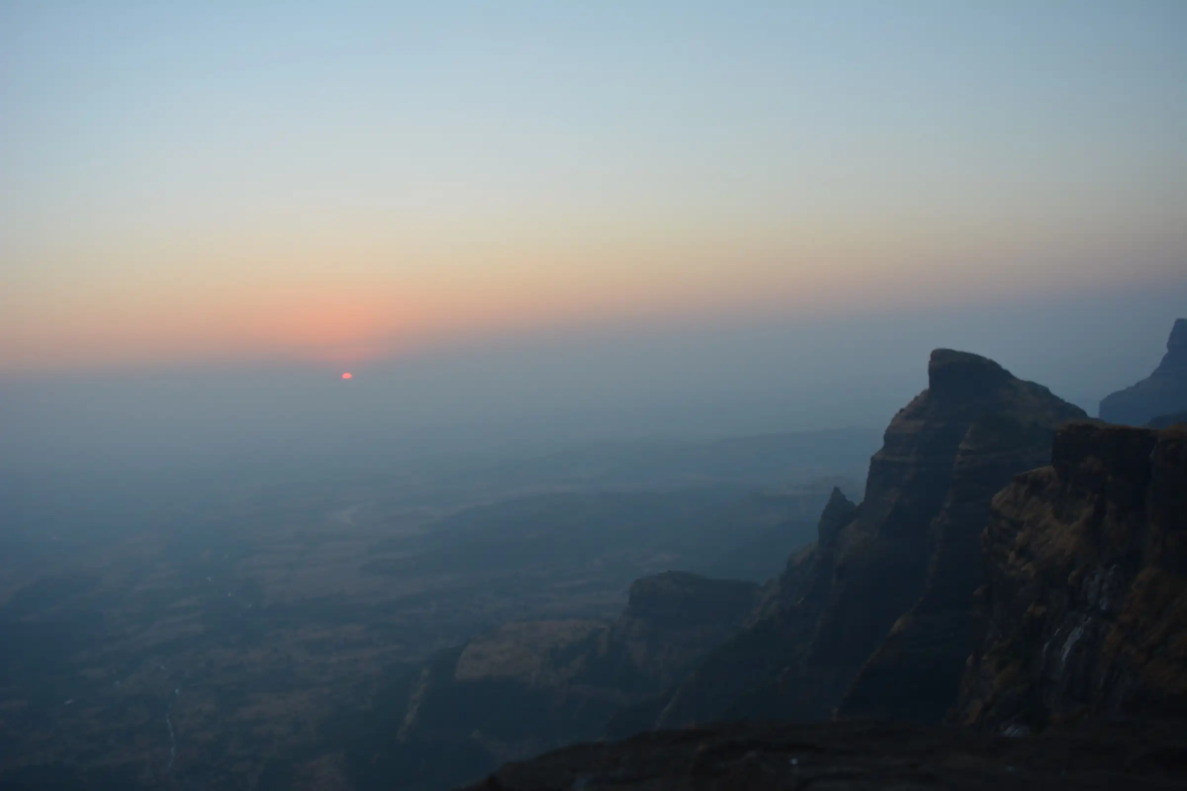 Sun setting into haze beyond the sheer rock walls of Konkan Kada on Harishchandragad