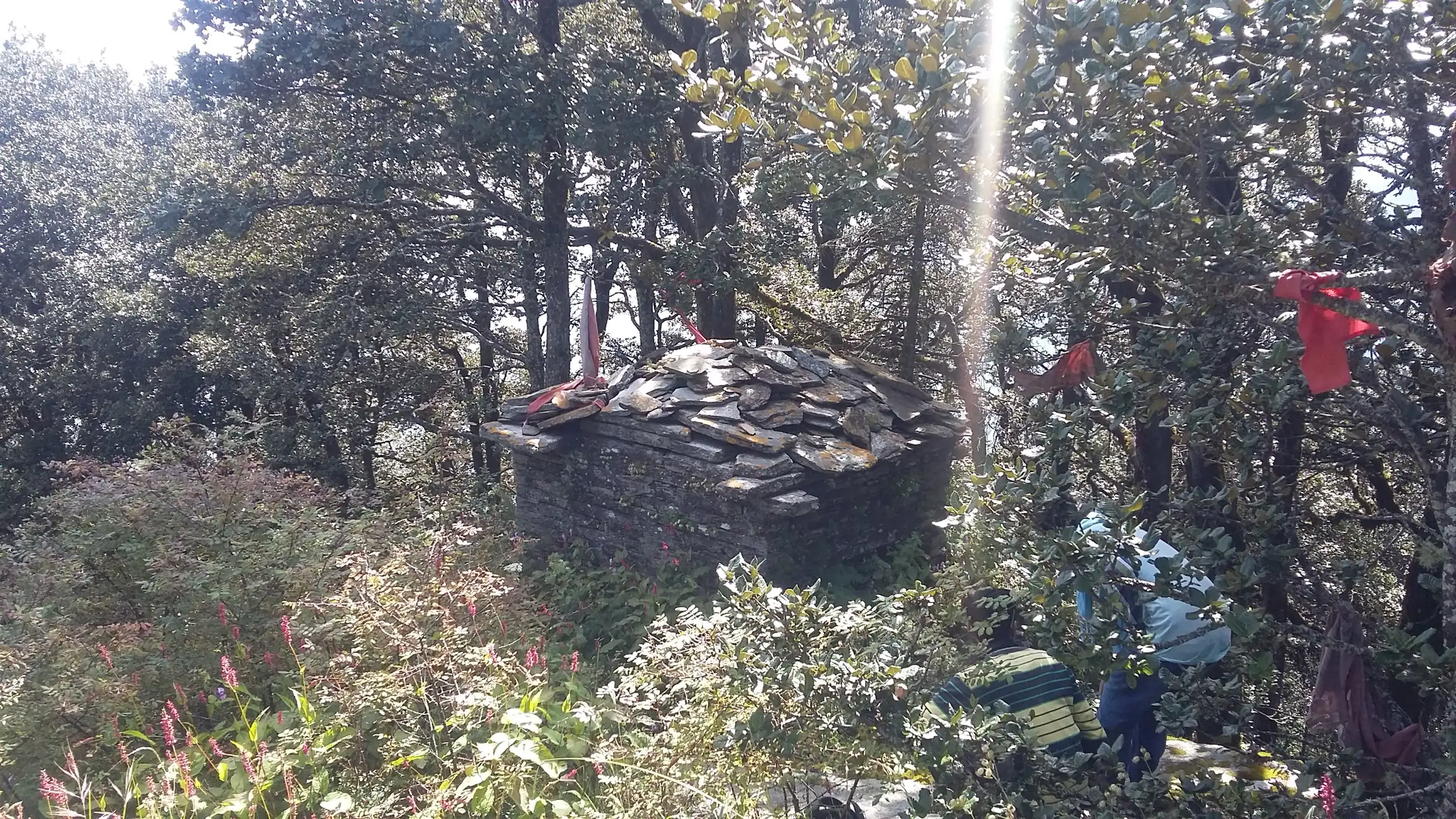 A small slate-roofed stone shrine with red cloth offerings in the forest at Kupad, Giri Ganga, with trekkers resting beside it