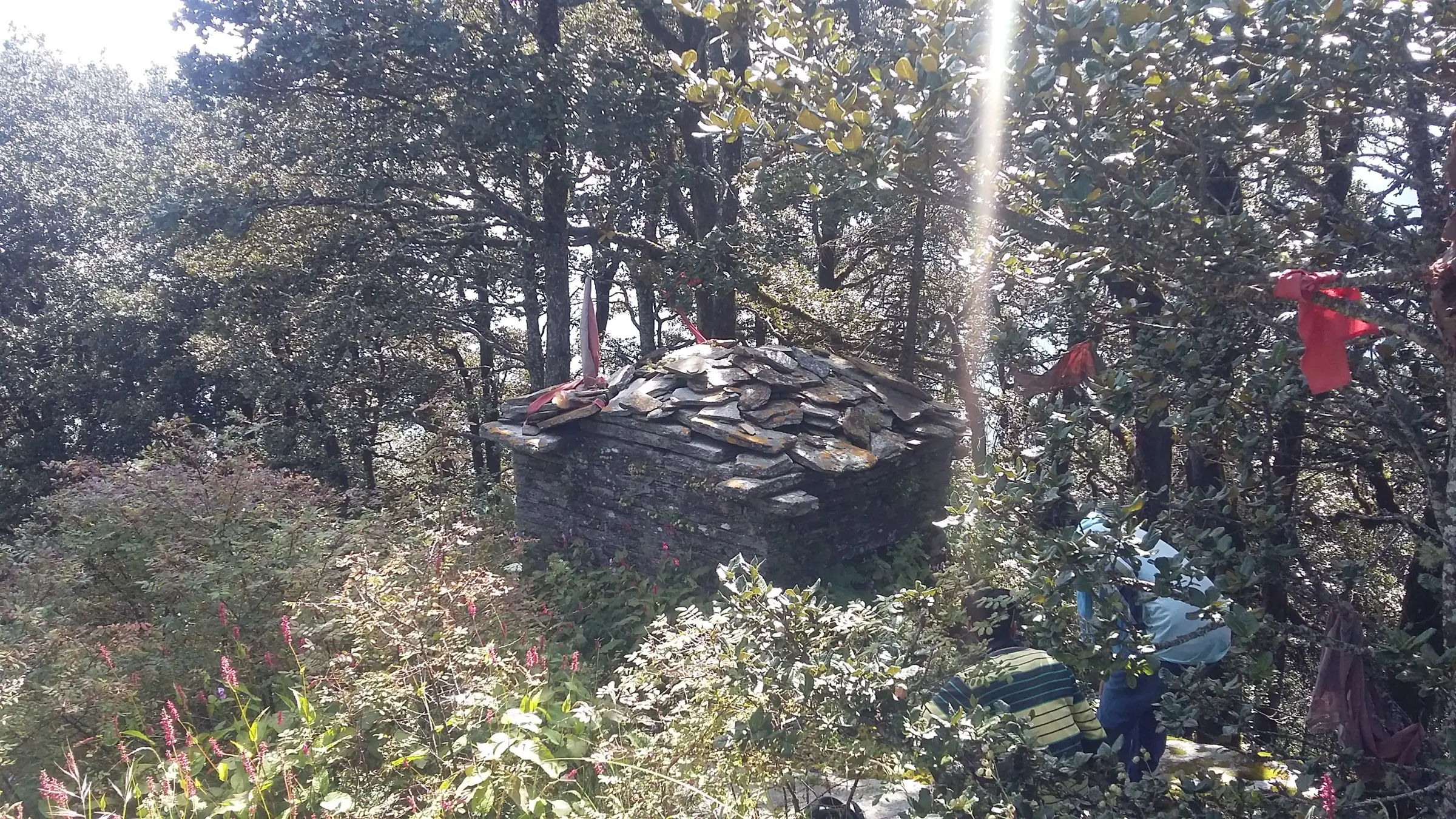 A small slate-roofed stone shrine with red cloth offerings in the forest at Kupad, Giri Ganga, with trekkers resting beside it
